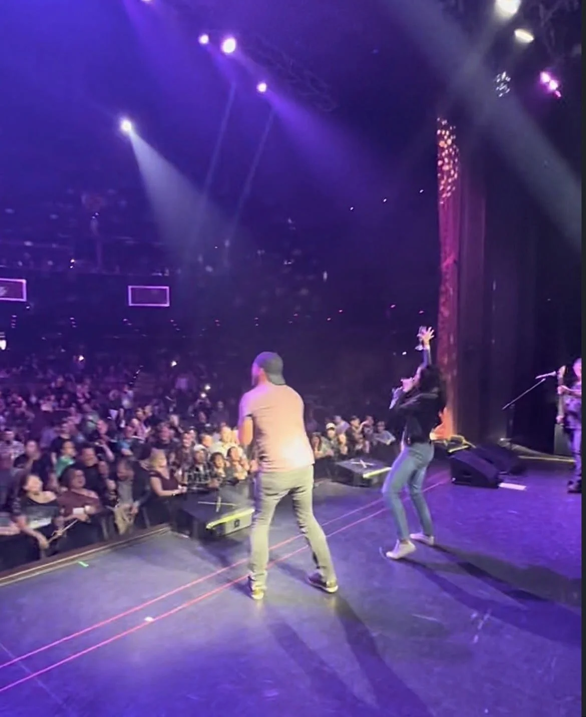 Performers on stage during a concert or event with a large audience in an indoor venue, stage lighting, and a curtain on the right side.
