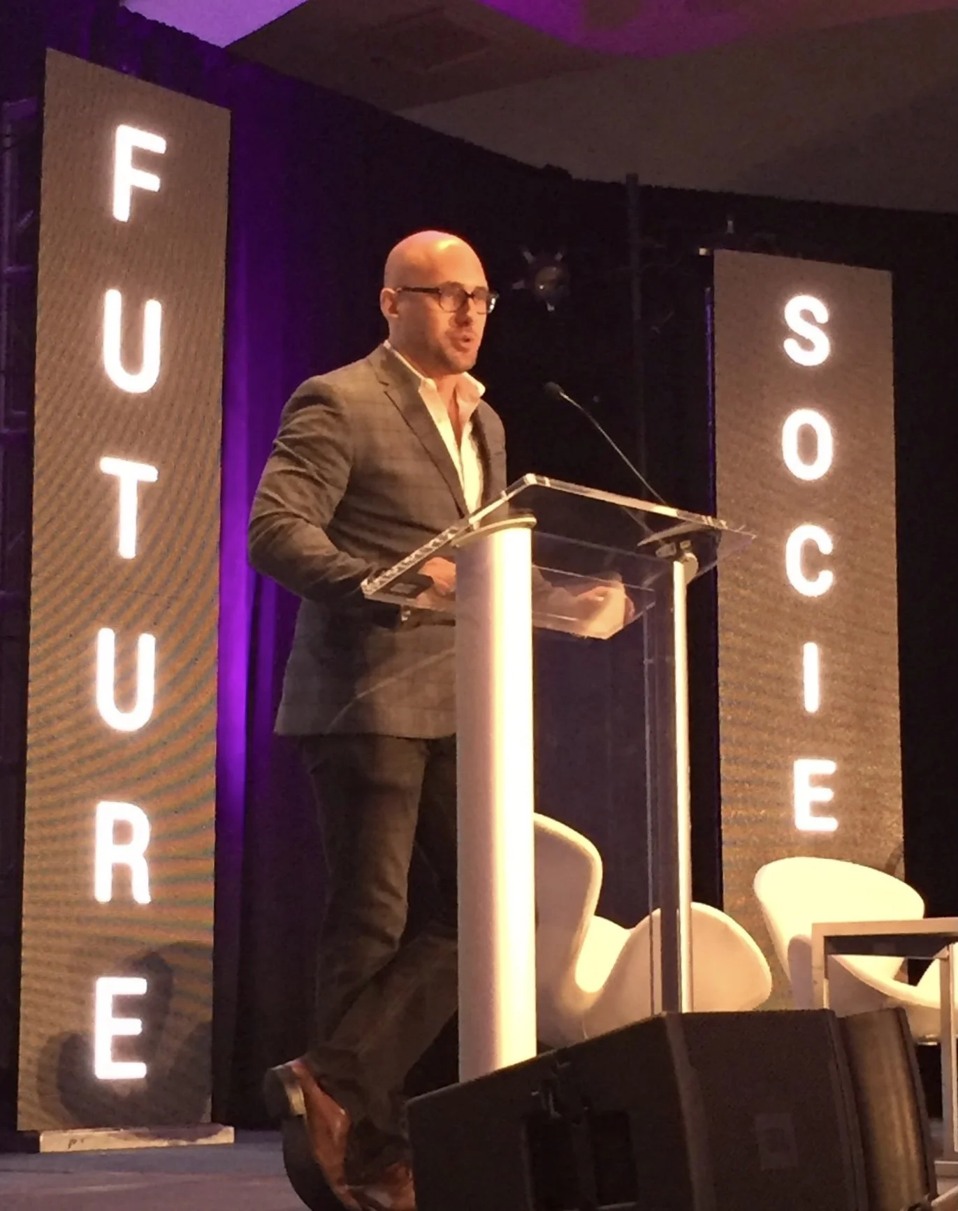 A man in a suit and glasses speaking at a podium on a stage with a microphone. Behind him are two tall vertical screens with the words 'FUTURE' and 'SOCiety' in bright letters.