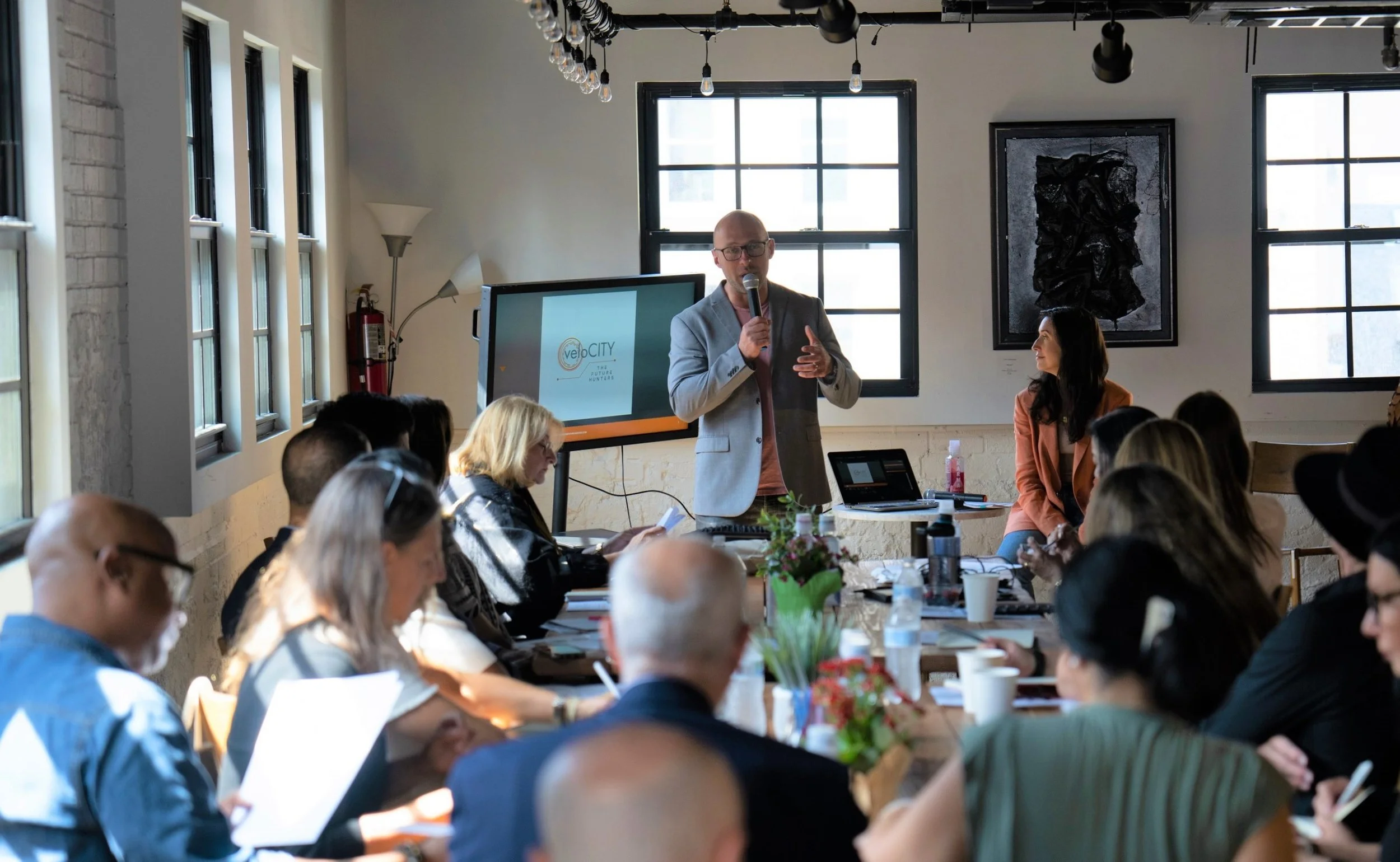 A man in a light gray blazer and glasses holding a microphone giving a presentation to an audience in a bright, modern room with large windows, black window frames, and an abstract black artwork on the wall.