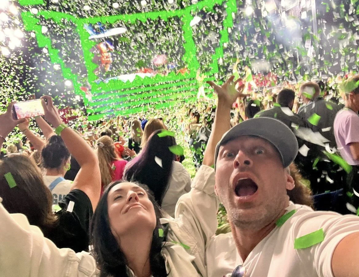 A crowd of people at a large event or concert, with some taking selfies and others cheering, surrounded by green and white confetti falling from above.