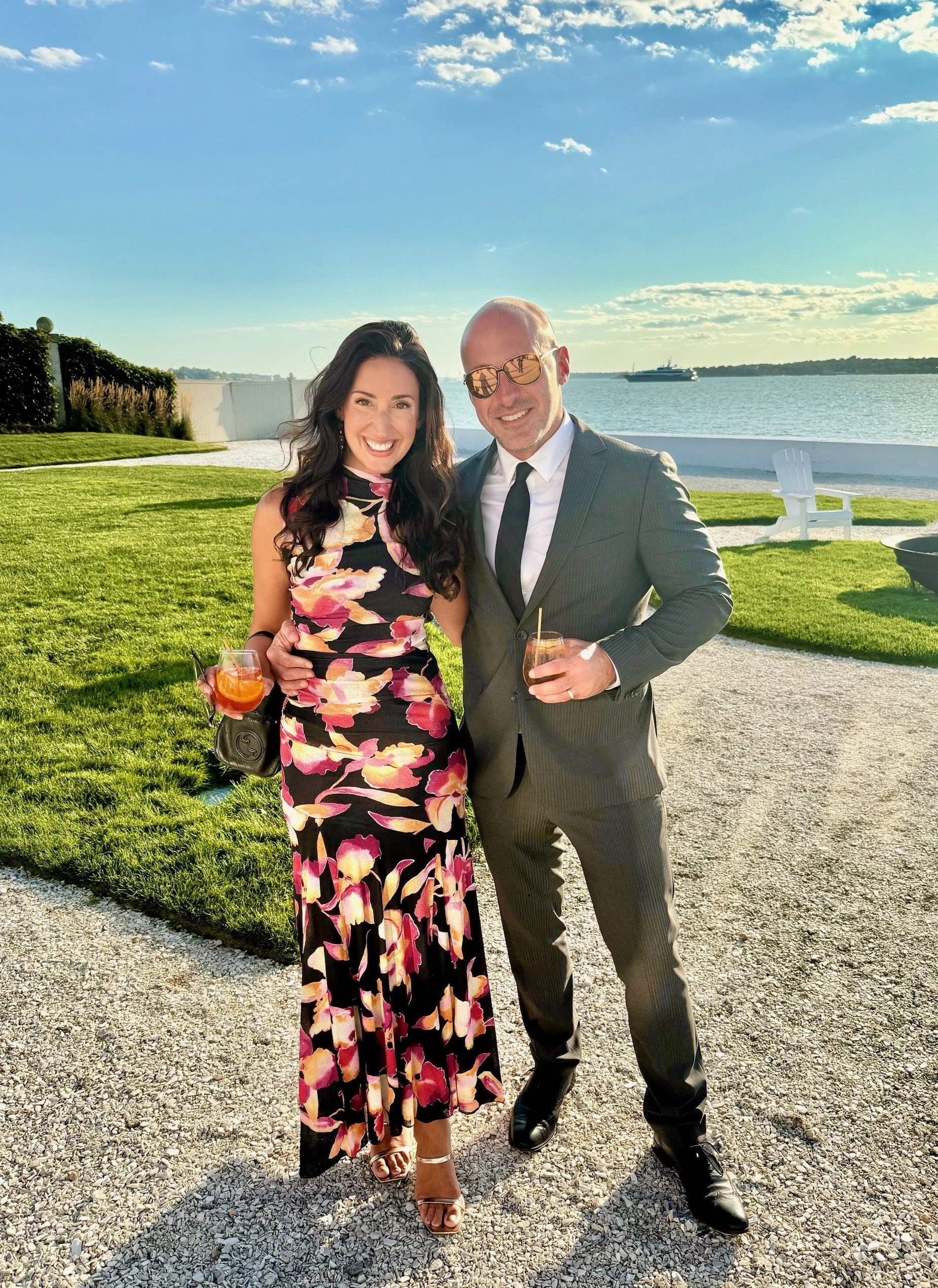 A smiling woman in a floral sleeveless dress and a man in a suit with sunglasses holding drinks, standing on a gravel path near the water with a grassy area and benches in the background, under a partly cloudy sky.