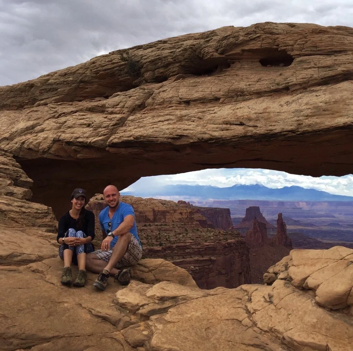 Two people sitting on a rock formation beneath a large natural rock arch in a desert landscape.