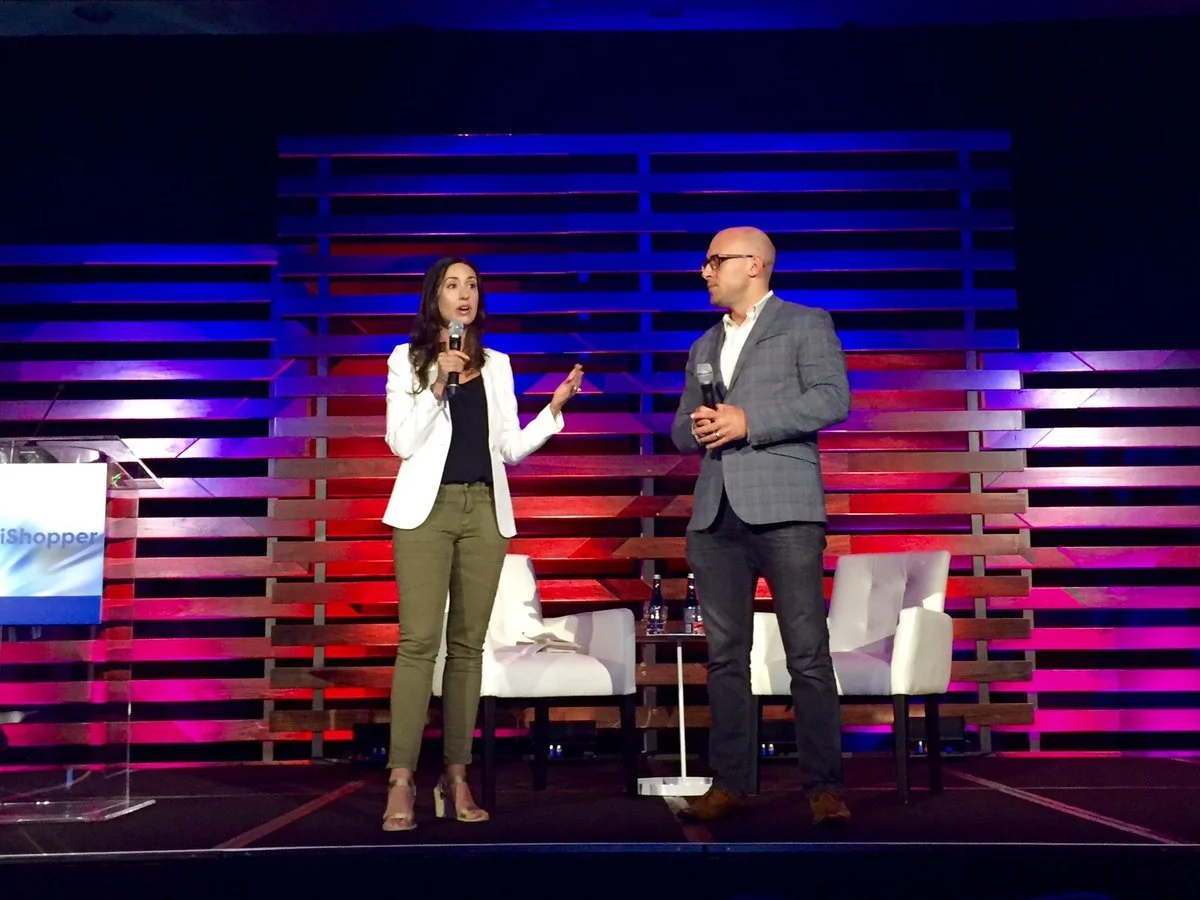 A woman and a man speaking on stage during a panel discussion or presentation. The woman has long dark hair and is wearing a white blazer, black top, and olive green pants. The man is bald, wearing glasses, a gray checkered blazer, and dark pants. They are holding microphones. There are two chairs and a small table with water bottles in the background, with colorful lighting behind them.