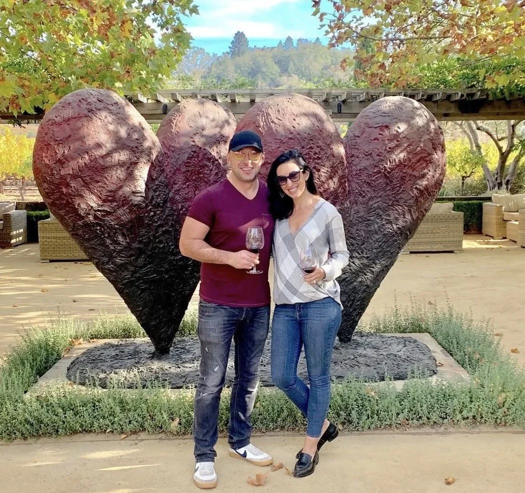 A smiling man and woman holding glasses of red wine stand in front of a large heart-shaped sculpture outdoors during autumn.