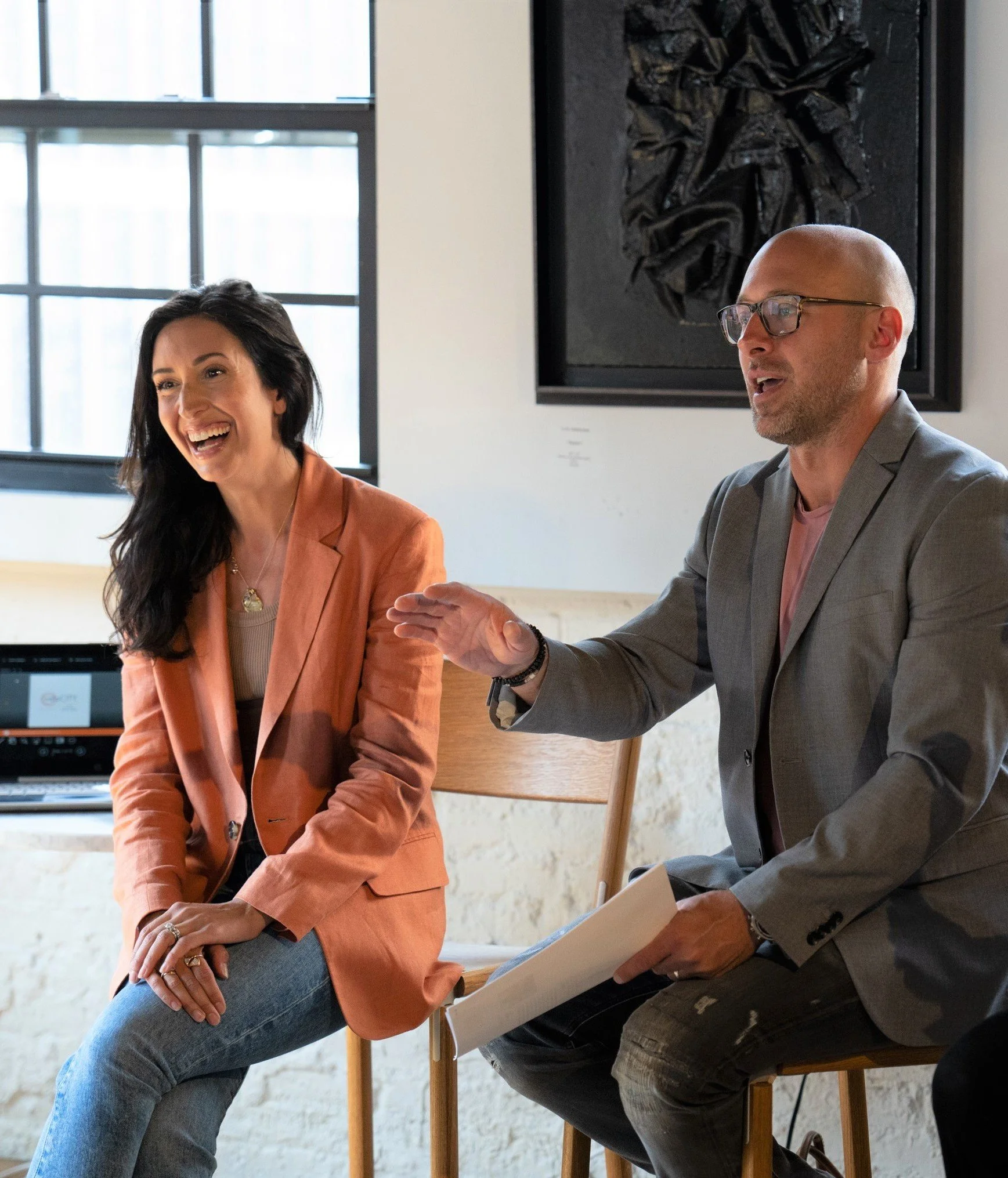 Two people, a woman with long black hair and a man with glasses and a shaved head, are sitting at a table in a bright room, engaged in conversation. The woman is smiling and wearing a peach-colored blazer, while the man is gesturing with his hand and holding a folder. There is a large window with grid panes behind them and a piece of black artwork on the wall.