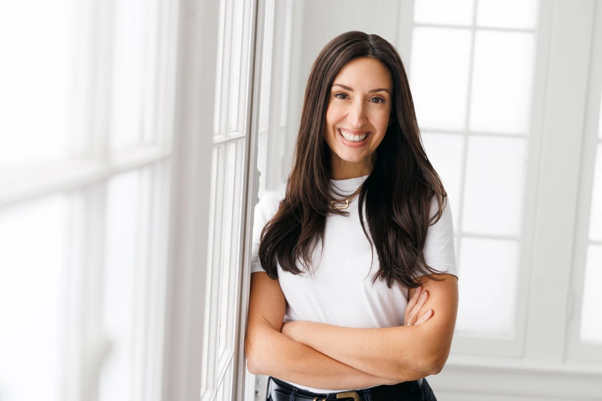 A woman with long dark hair, smiling, standing with arms crossed inside a bright room with large white-framed windows.