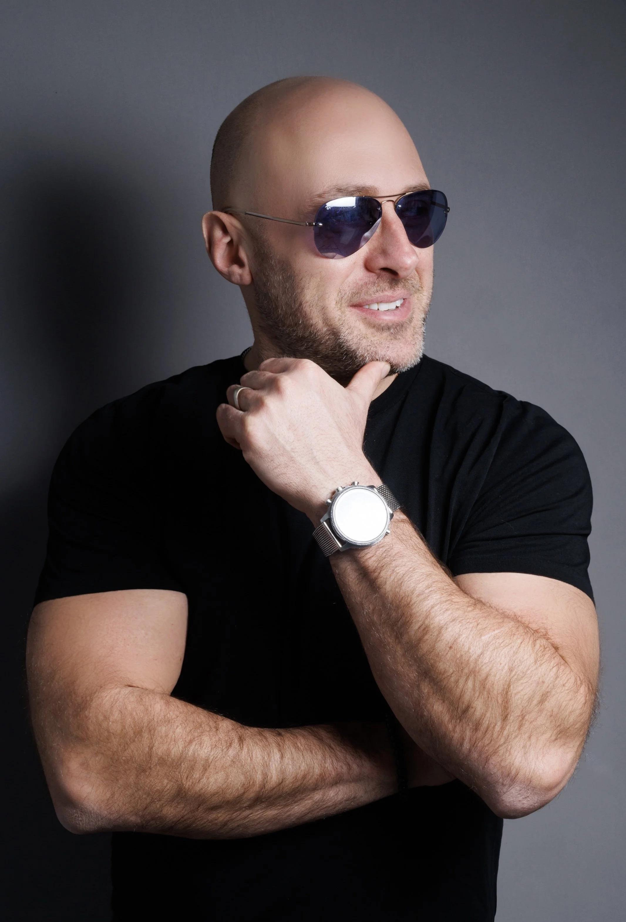A man with a bald head, sunglasses, and a beard, wearing a black t-shirt, is smiling slightly while resting his chin on his hand. He is also wearing a watch on his left wrist.