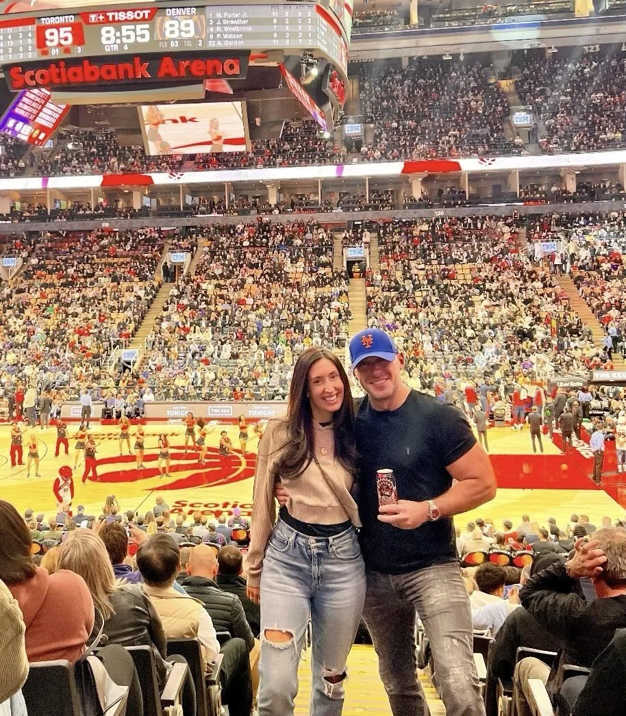Two people smiling at a basketball game in an arena with a large crowd. The woman has long dark hair and is wearing a beige sweater and ripped jeans. The man is wearing a blue cap, black t-shirt, and holding a drink. The arena is filled with spectators, and the game is in progress with the score displayed at the top.