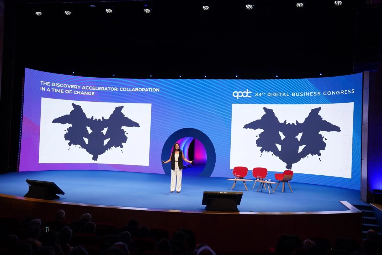 A woman is standing on stage at the 34th Digital Business Congress, presenting with a large screen behind her displaying two black and white symmetrical abstract images with labels, 'The Discovery Accelerator: Collaboration in a Time of Change'. Ther