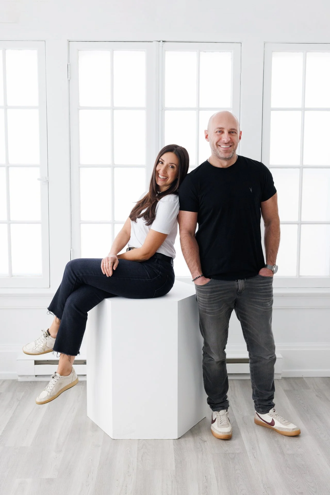 A woman sitting on a white platform smiling, while a man stands next to her, both indoors in front of large windows, wearing casual clothes.