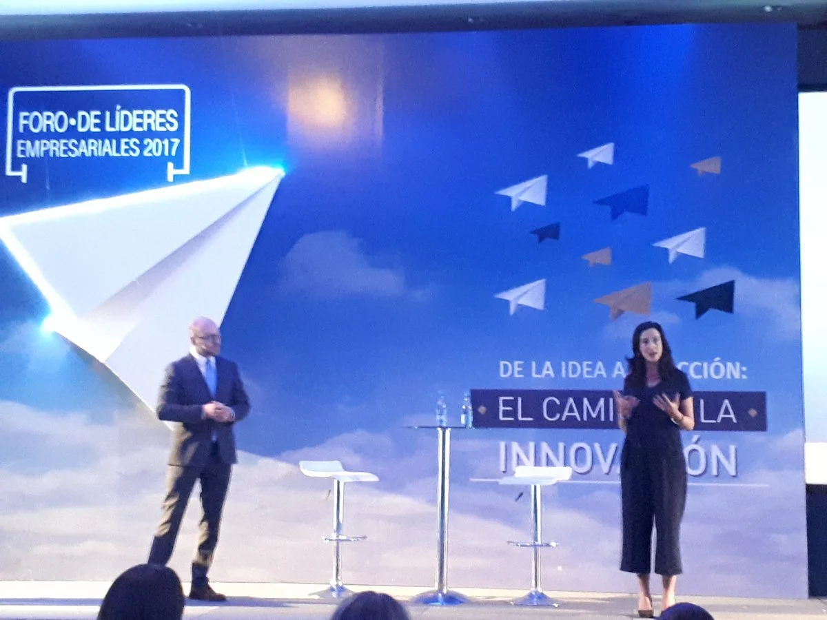 Two speakers on stage at the 2017 Business Leaders Forum, with a backdrop featuring paper airplane graphics and the text in Spanish about transforming ideas into action through innovation.
