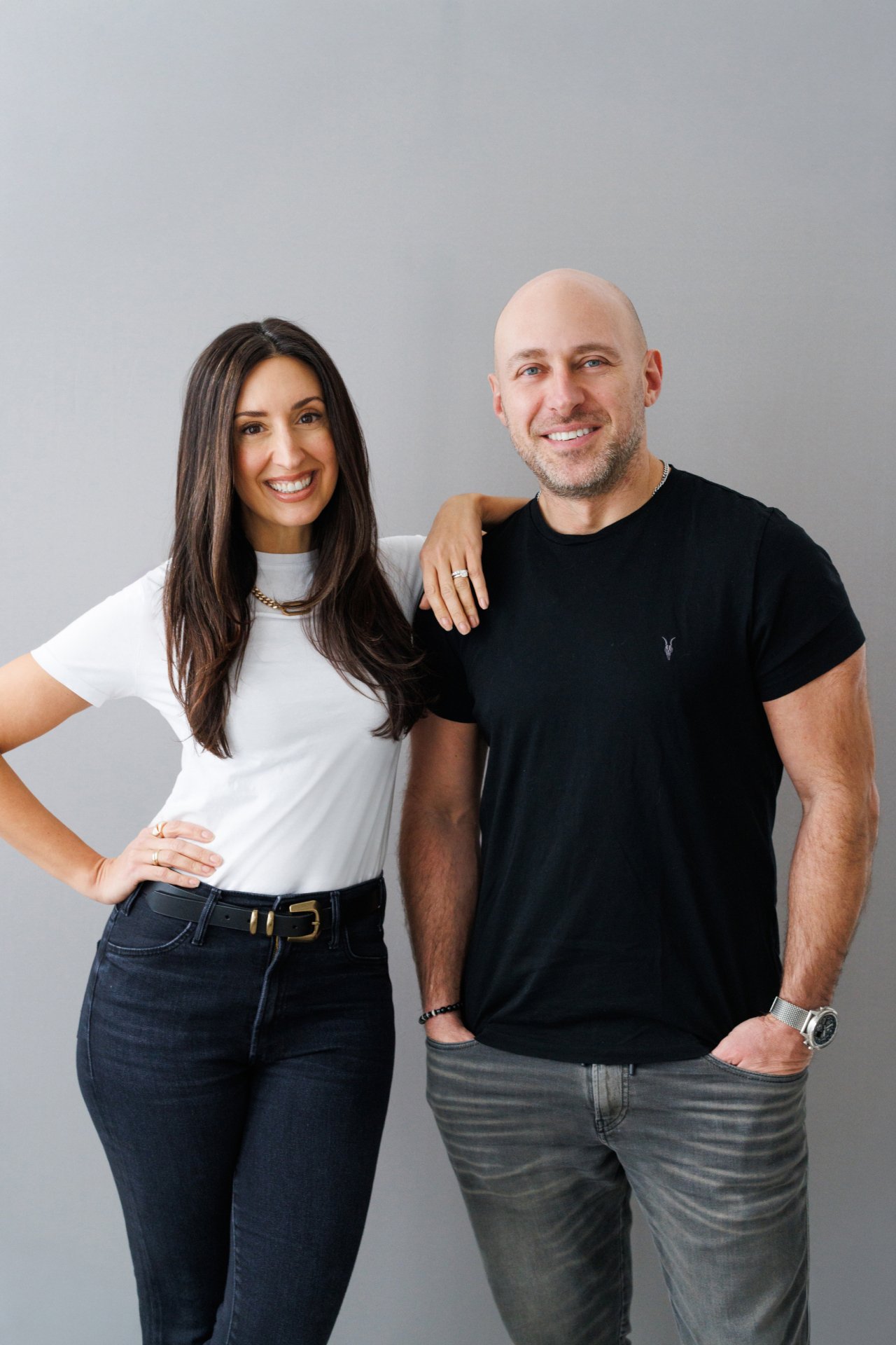 A smiling woman with long dark hair wearing a white t-shirt and dark jeans with a black belt, standing next to a smiling bald man with a beard wearing a black t-shirt and gray jeans, against a plain gray background.