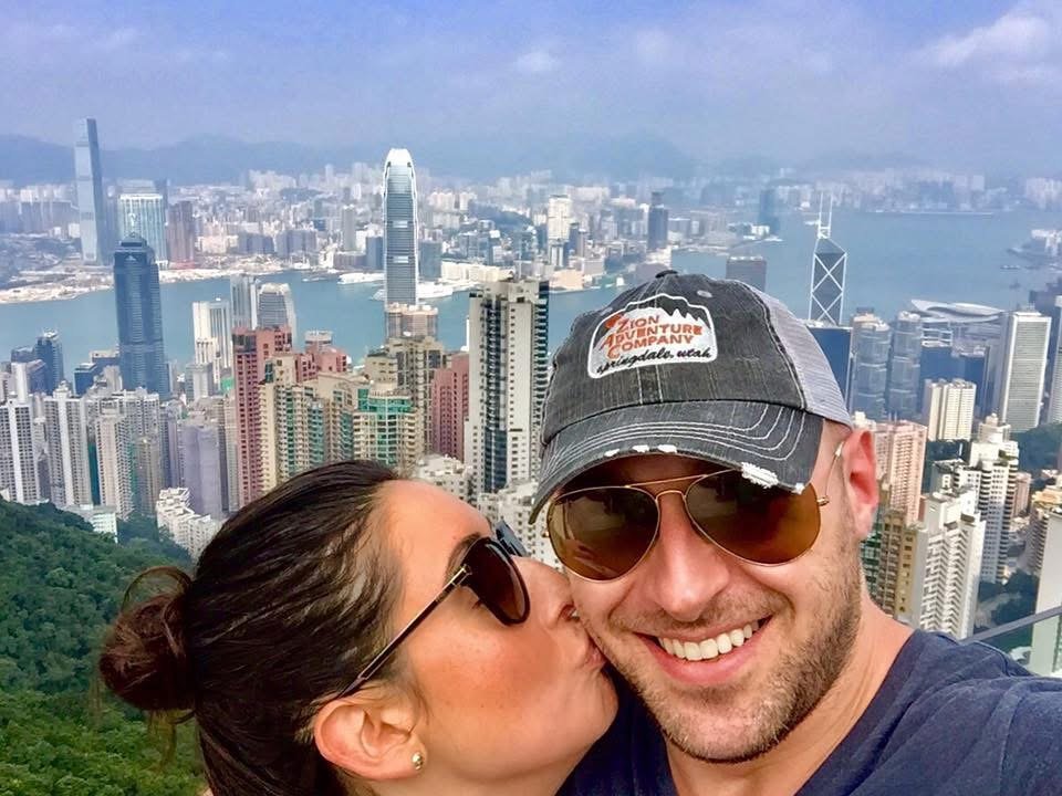 A couple taking a selfie with a city skyline and harbor in the background.