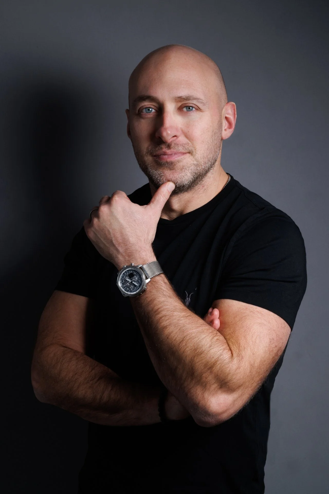 A bald man with blue eyes and a beard, wearing a black t-shirt, is posing with his right hand touching his chin and his left arm crossed over his chest, standing against a dark background.