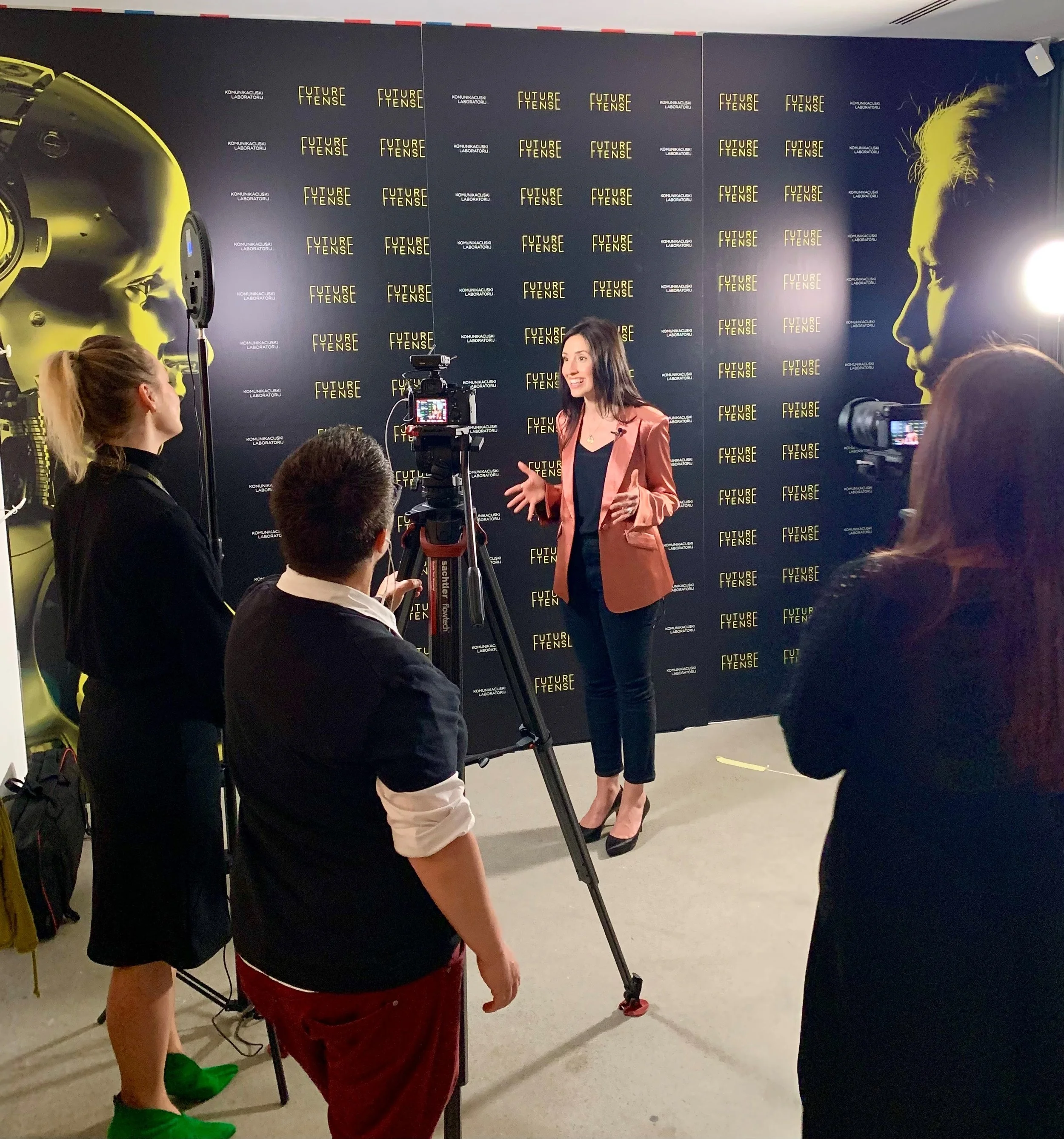 A woman in a pink blazer and black top speaking in front of a black background with yellow text reading 'Future Tense' and 'KOMUNIKACJSKI LABORATORIJ.' Several people are in the audience, including a woman with long curly hair and a black blazer, a p