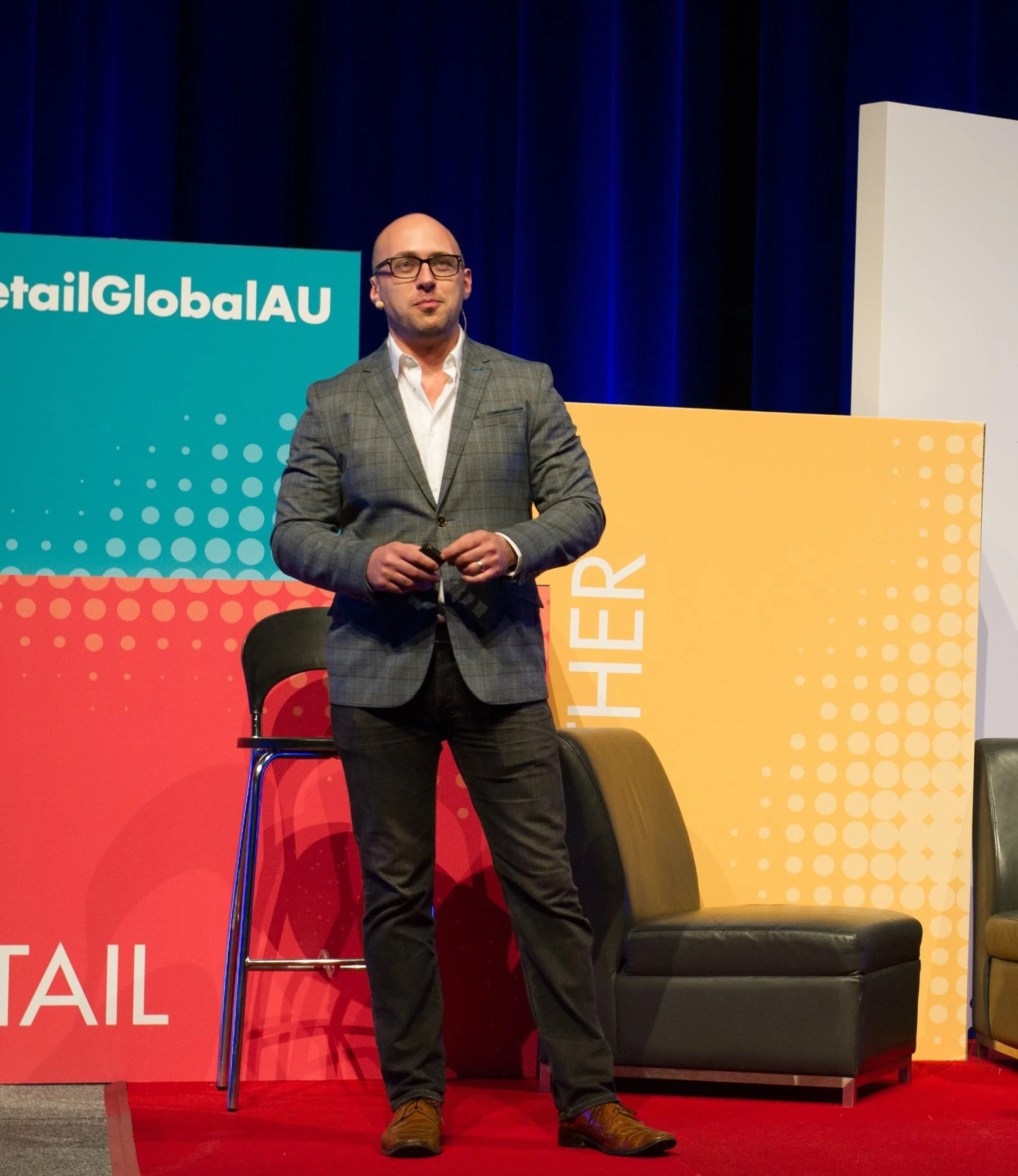 A man wearing glasses, a gray plaid suit jacket, white shirt, dark pants, and brown boots stands on a red carpet at a conference or event. Behind him are colorful banners with text and dotted patterns, and a row of black chairs.