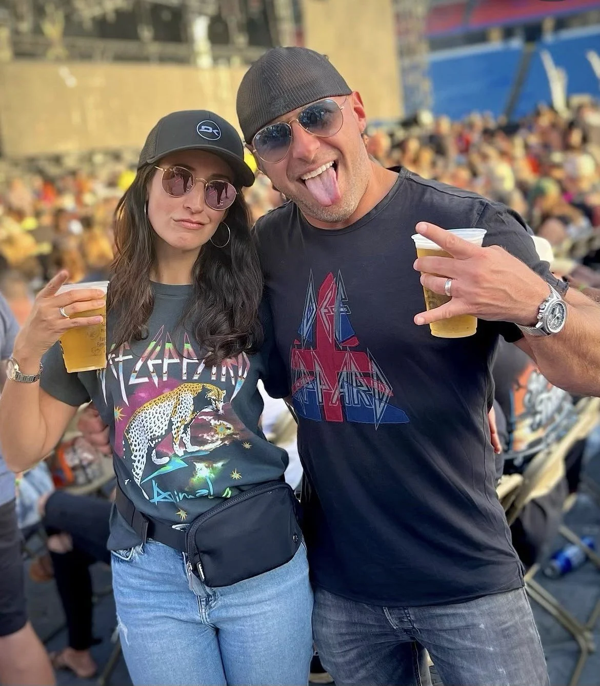 A man and woman at a crowd event, both wearing sunglasses and holding beer cups, smiling and making hand gestures. The woman wears a black cap, Metallica T-shirt, and casual jeans, while the man wears a dark cap, Grateful Dead T-shirt, and a large watch.