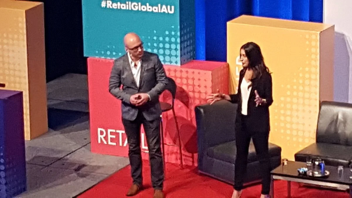 Two people speaking on stage at a conference with colored cube decorations and a blue screen background. The man is wearing a gray suit and glasses, and the woman is dressed in black with long dark hair.
