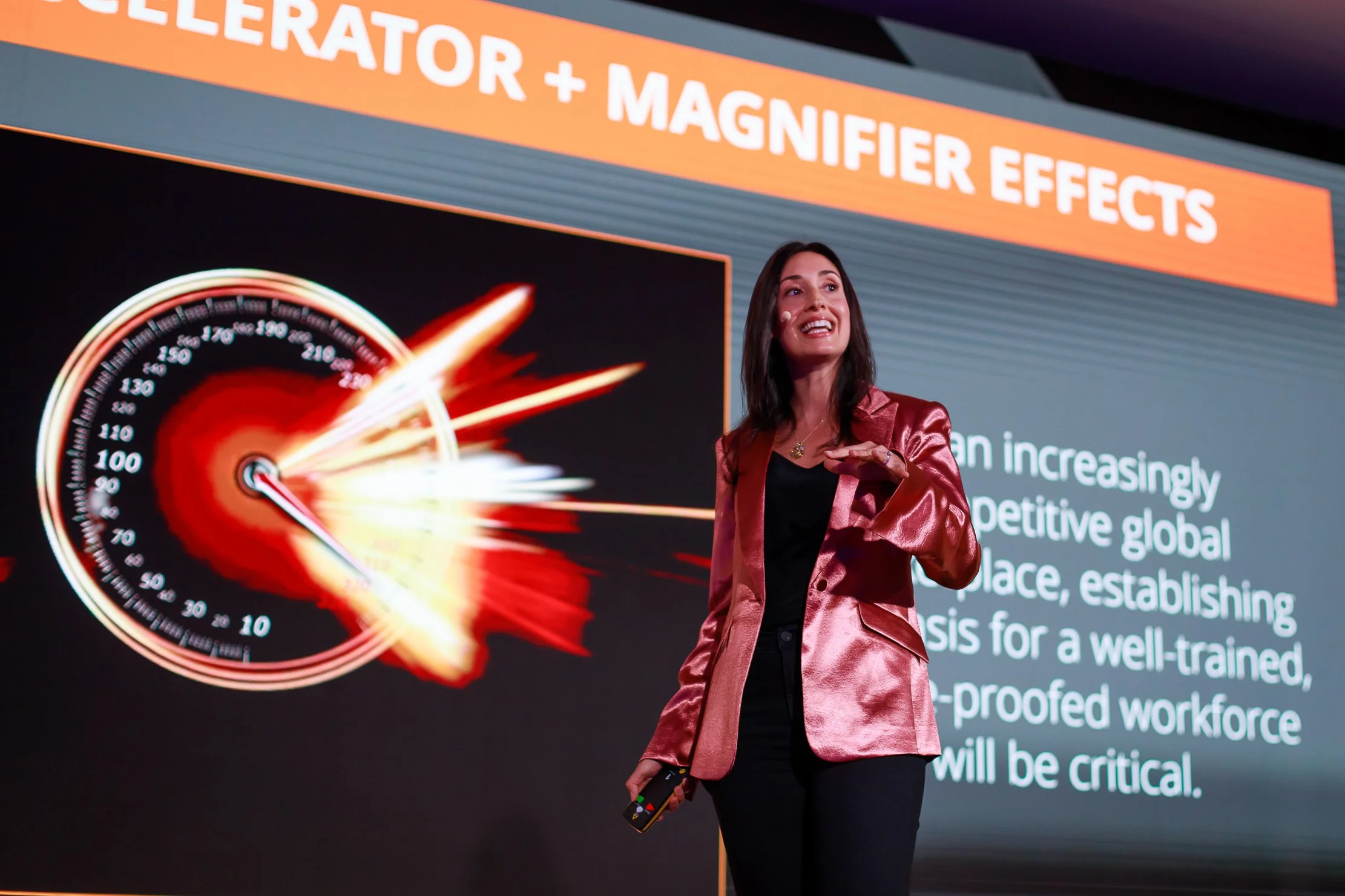 A woman in a pink jacket giving a presentation on effects of laser and magnet technology, with a large screen displaying a fiery graphic and some text behind her.