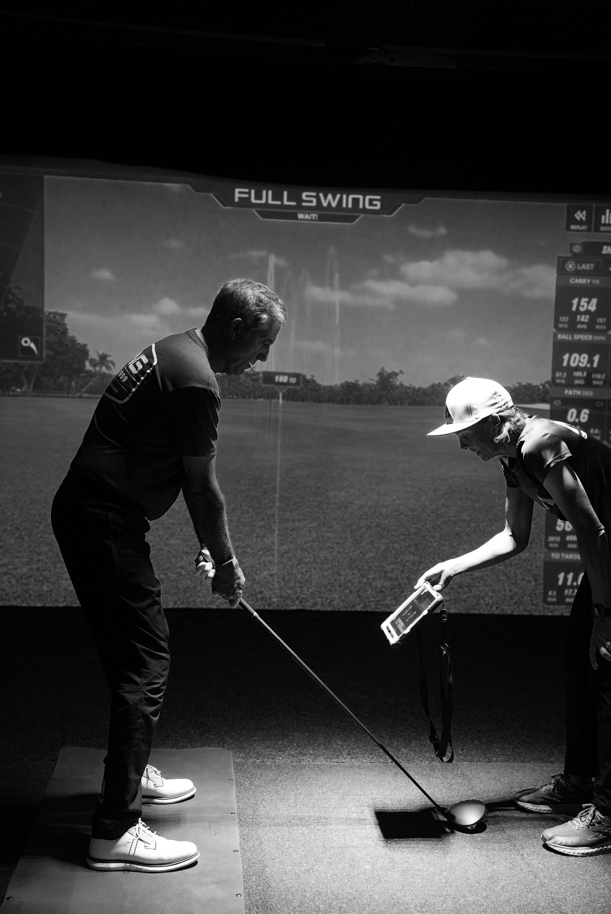 A man and a woman examining a golf club and ball in an indoor golf simulation room with a digital screen showing golf stats and a virtual golf course in the background.