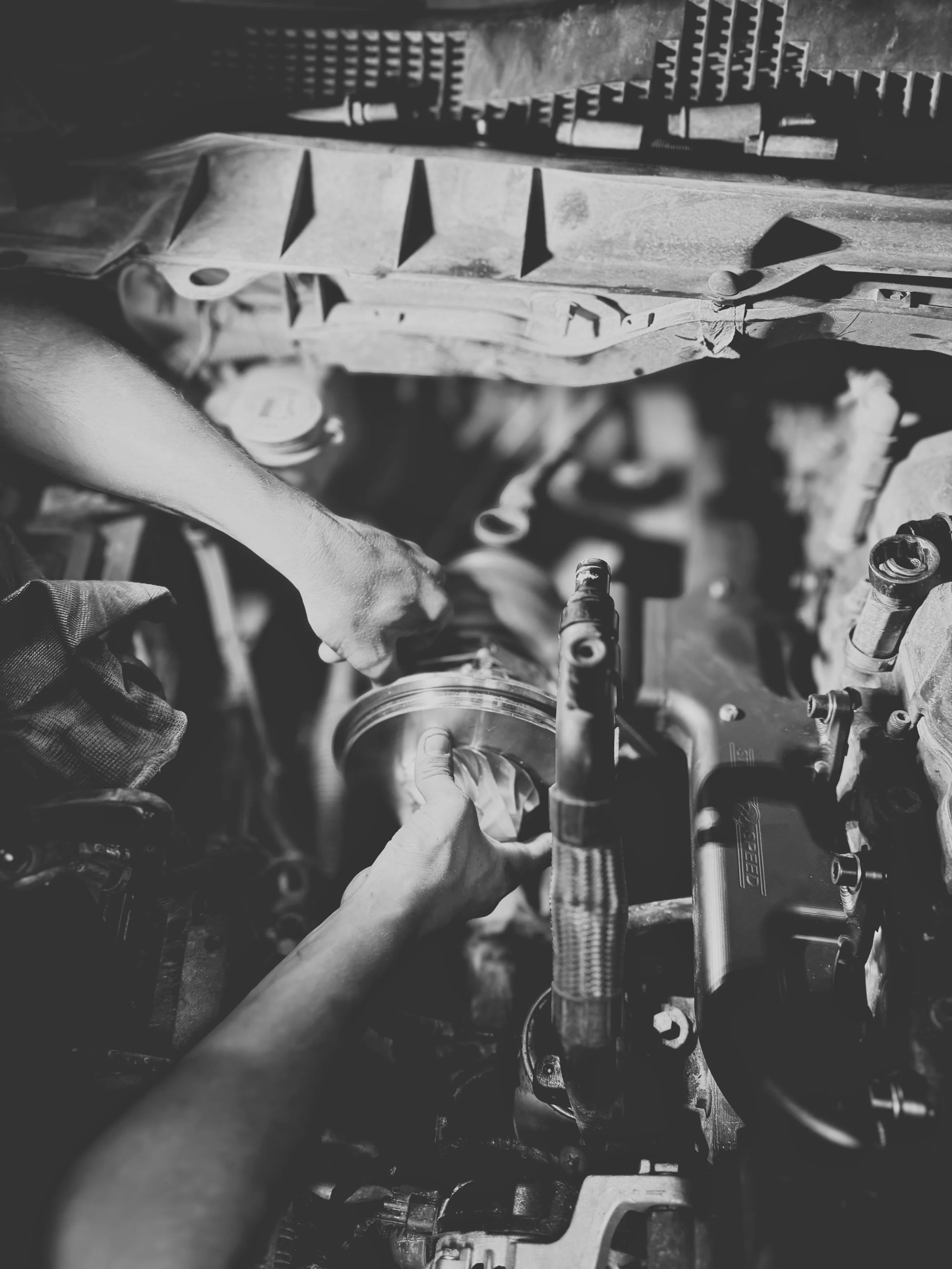 Two hands working on an engine in a car, with tools and engine parts visible, in black and white.