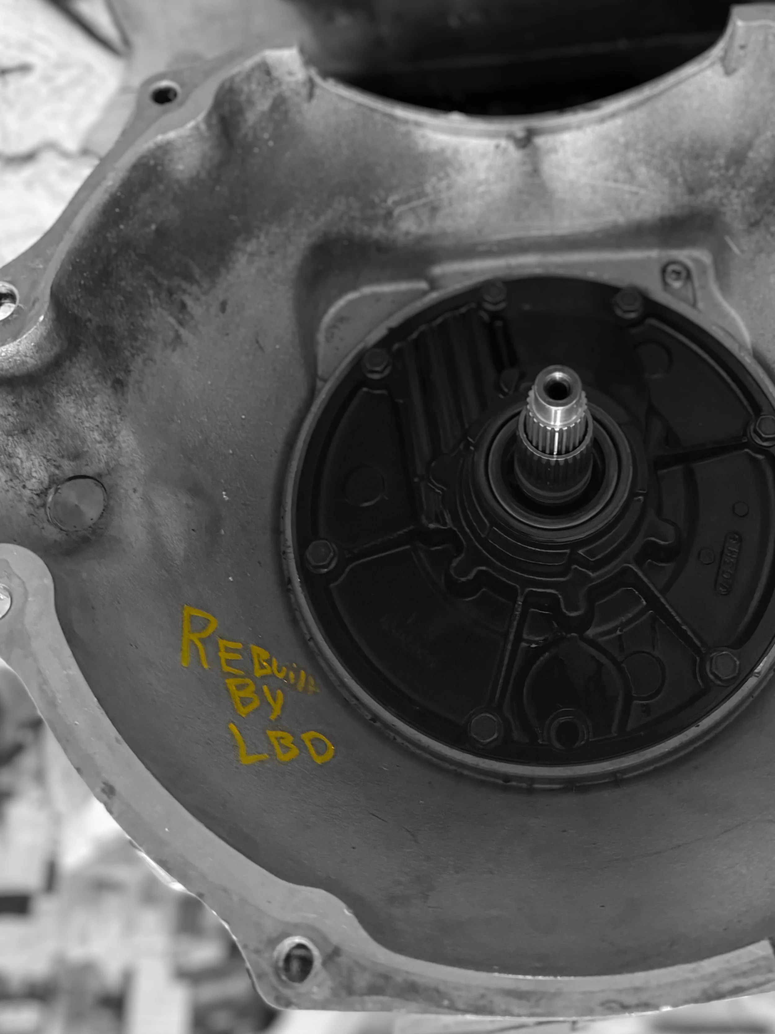 Close-up of a vehicle transmission component with yellow handwritten text 'Rebuilt by LBO' on the metal surface.