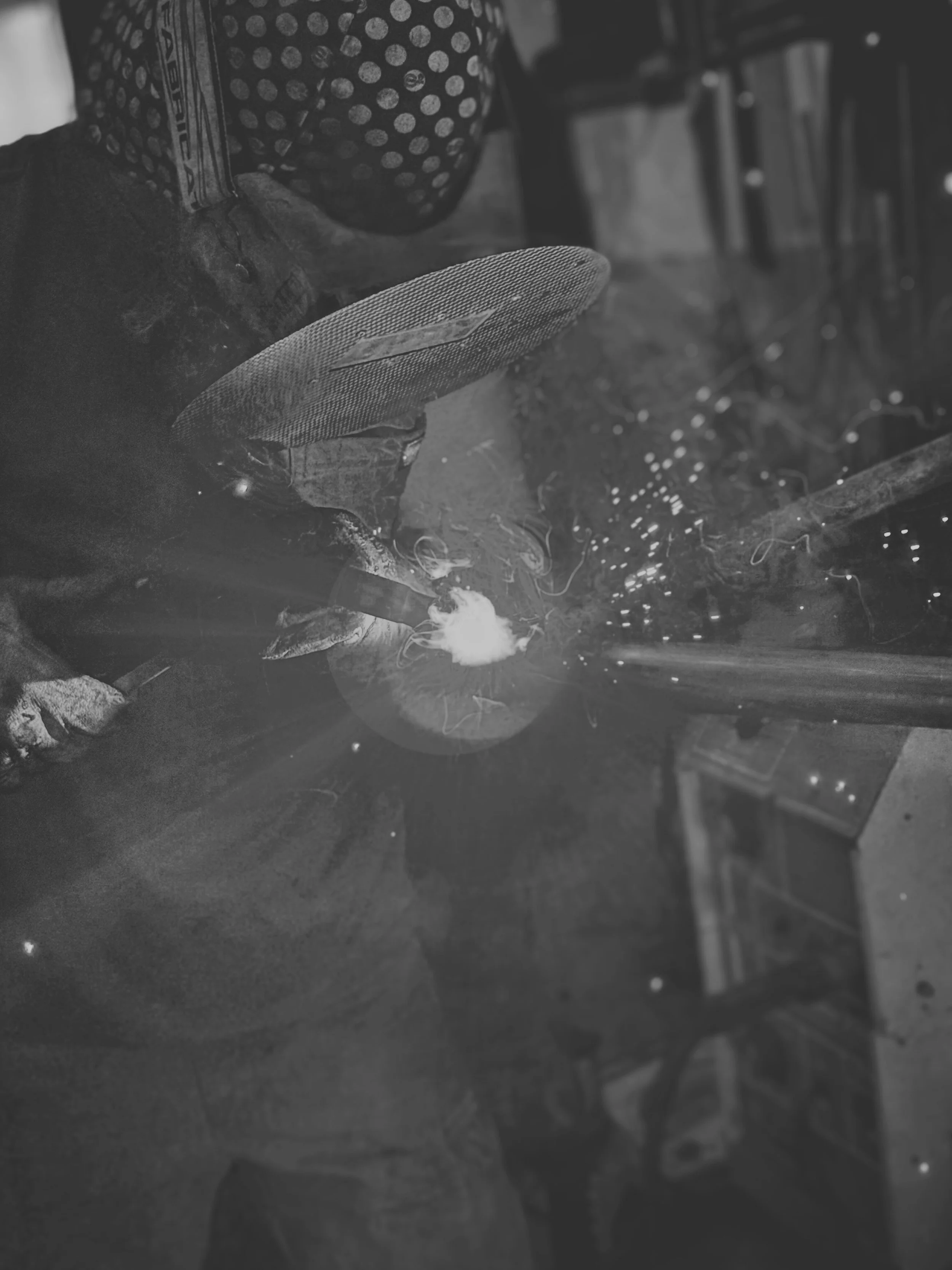 A person welding metal, sparks flying, wearing a polka dot face mask and protective gloves, in a workshop or industrial setting.