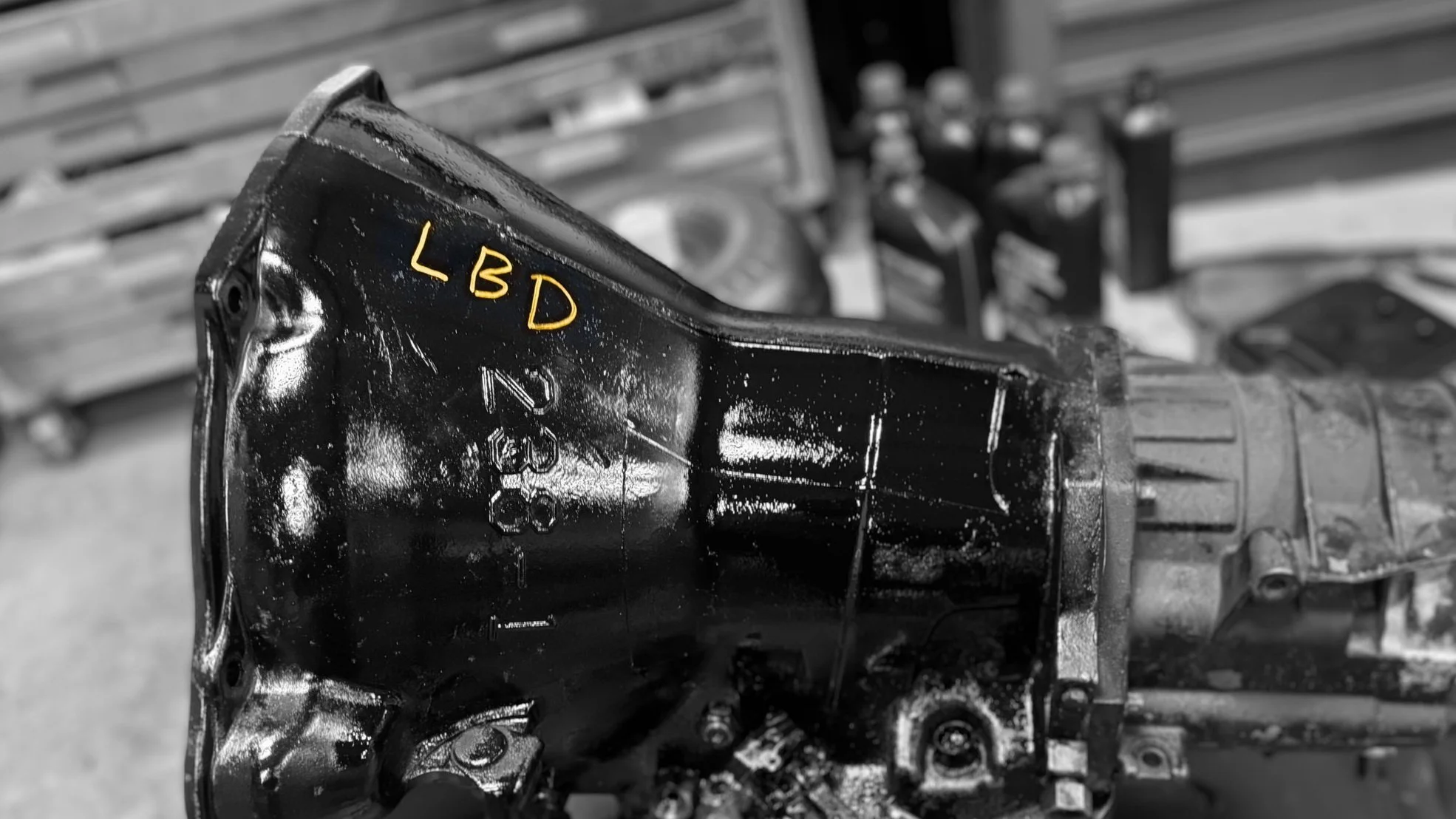 A close-up view of a black, greasy transmission with the handwritten yellow letters 'LBD' on it, in a workshop or garage setting.