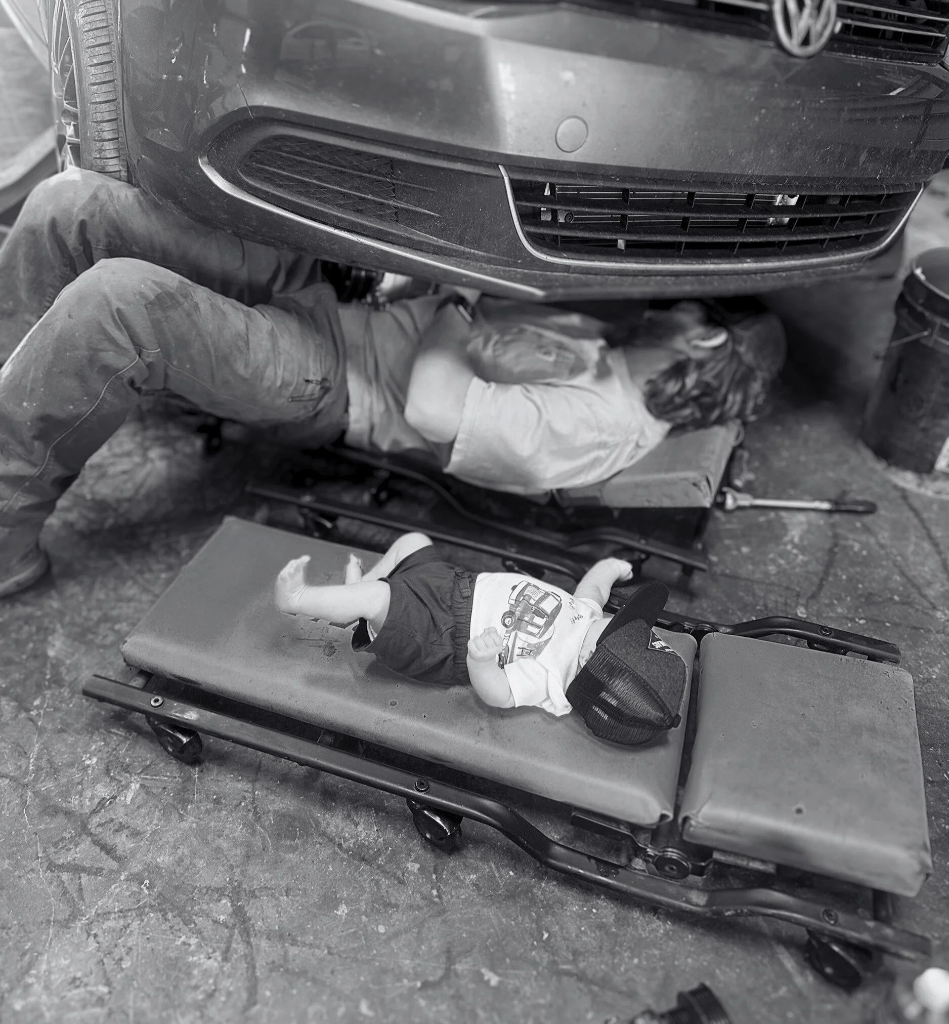 A mechanic working under a car on a creeper with a young child lying on a nearby wheeled stretcher.