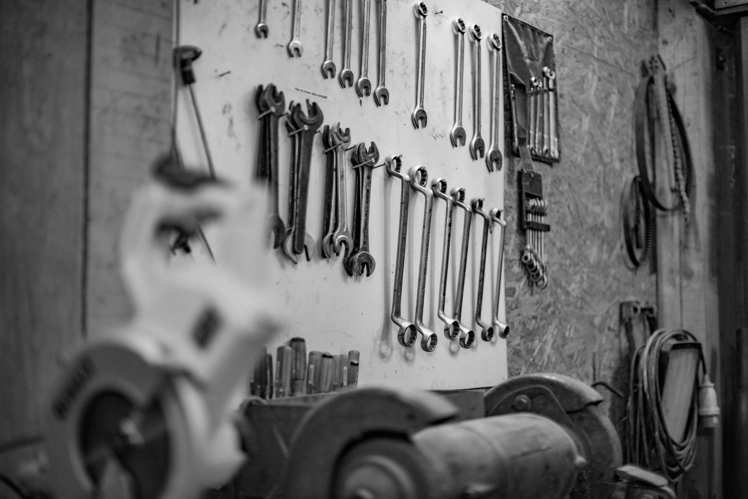 A black and white photo of a workshop wall with various metal tools hanging, including wrenches, pliers, and a saw, along with some other equipment and cords.