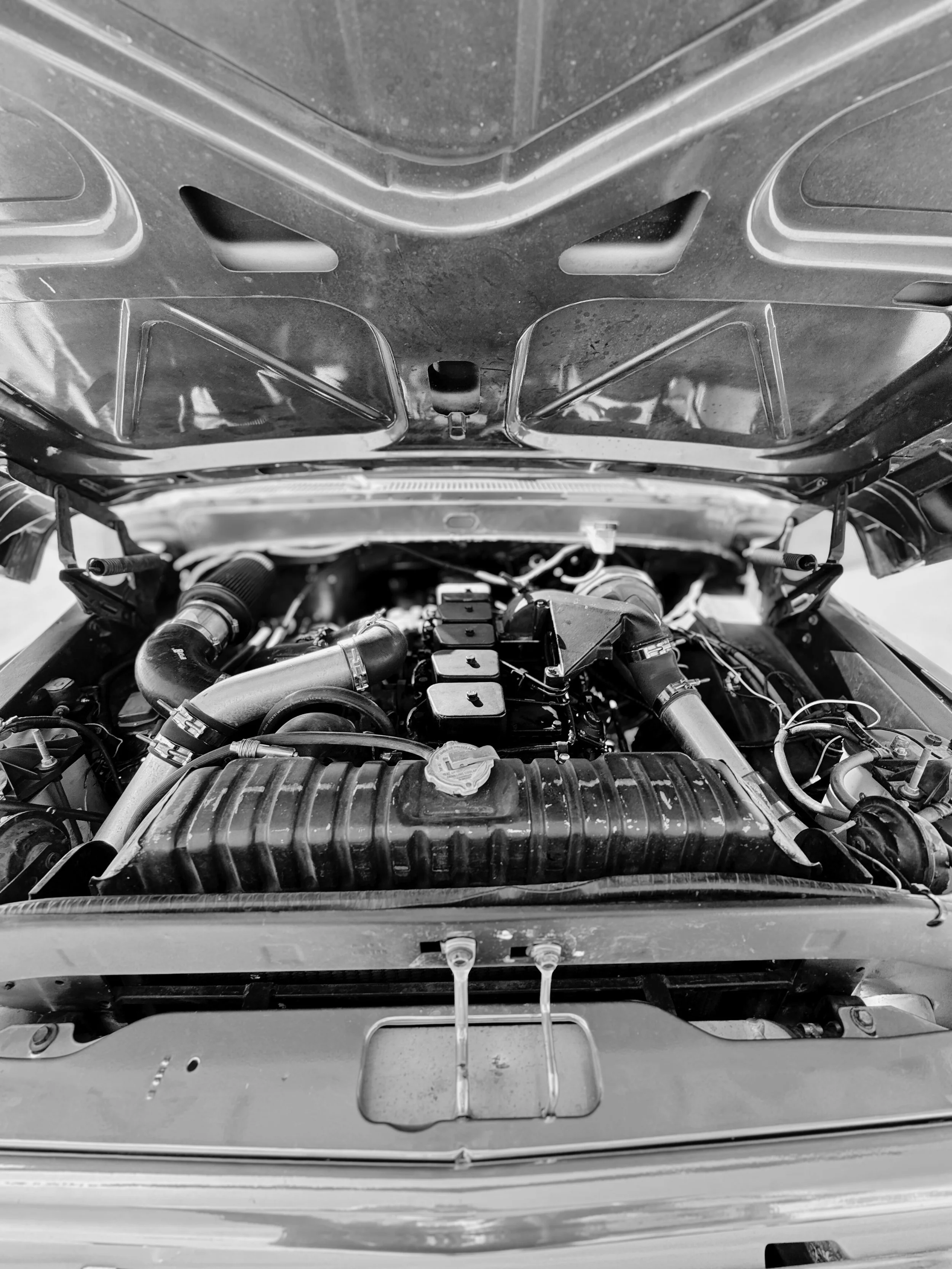 Black and white photo of a car engine with the hood open, showing various engine components and hoses.