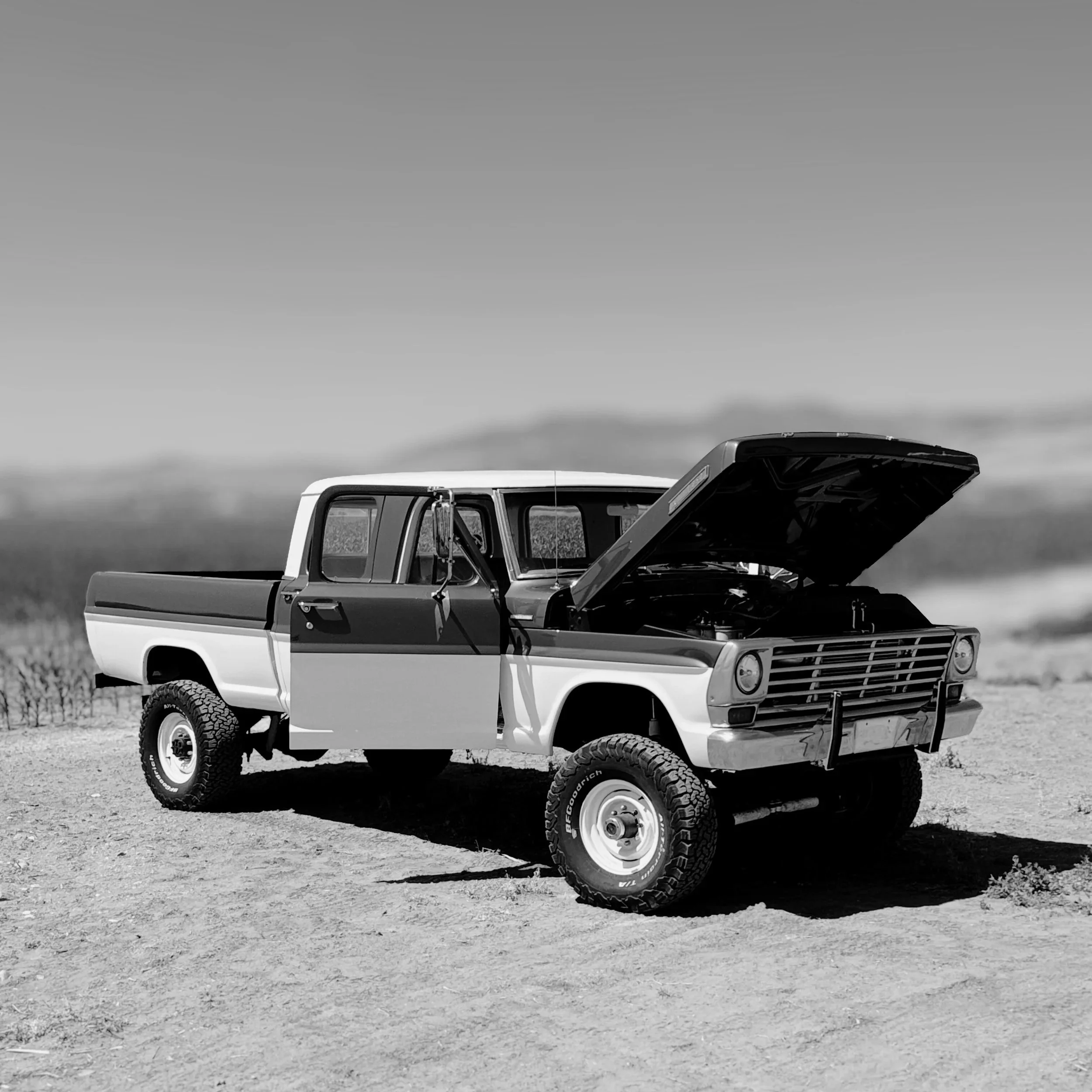 A black and white photo of a pickup truck parked on a dirt road with the hood open and the driver's side door open in a rural area with open sky and distant horizon.