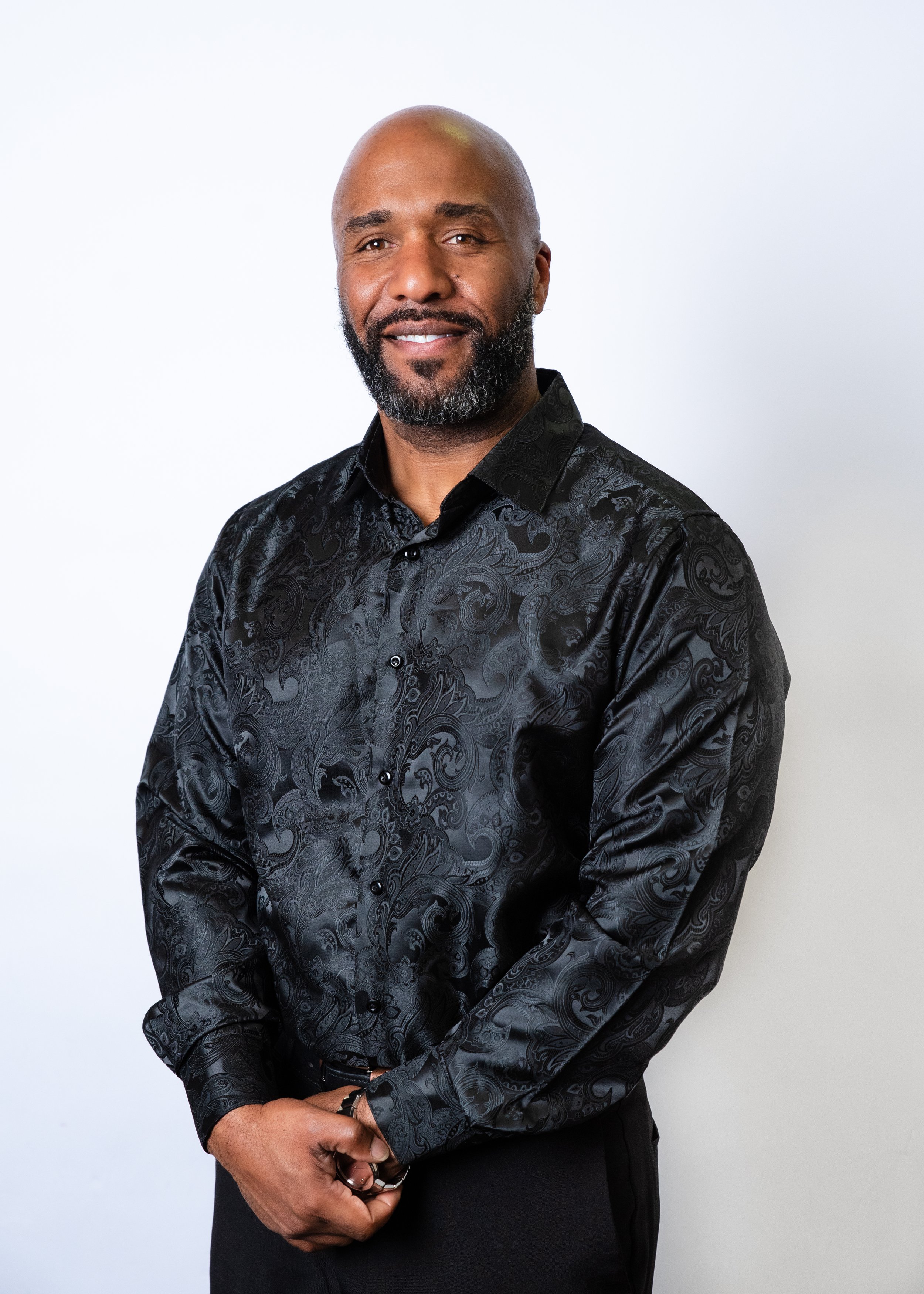 A man with a shaved head and beard wearing a black shirt against a peach-colored background.