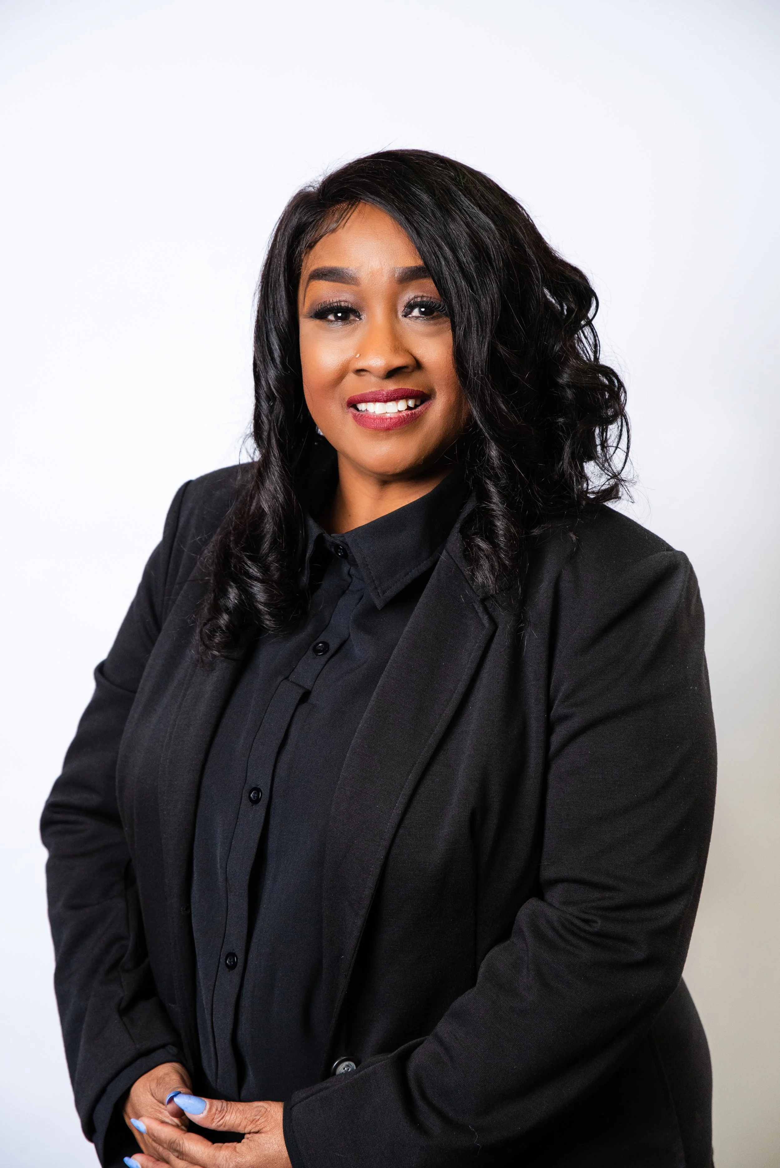 A woman with long, dark, curly hair, smiling, wearing a black shirt and a black scarf, standing against a gradient background from light peach to darker peach.