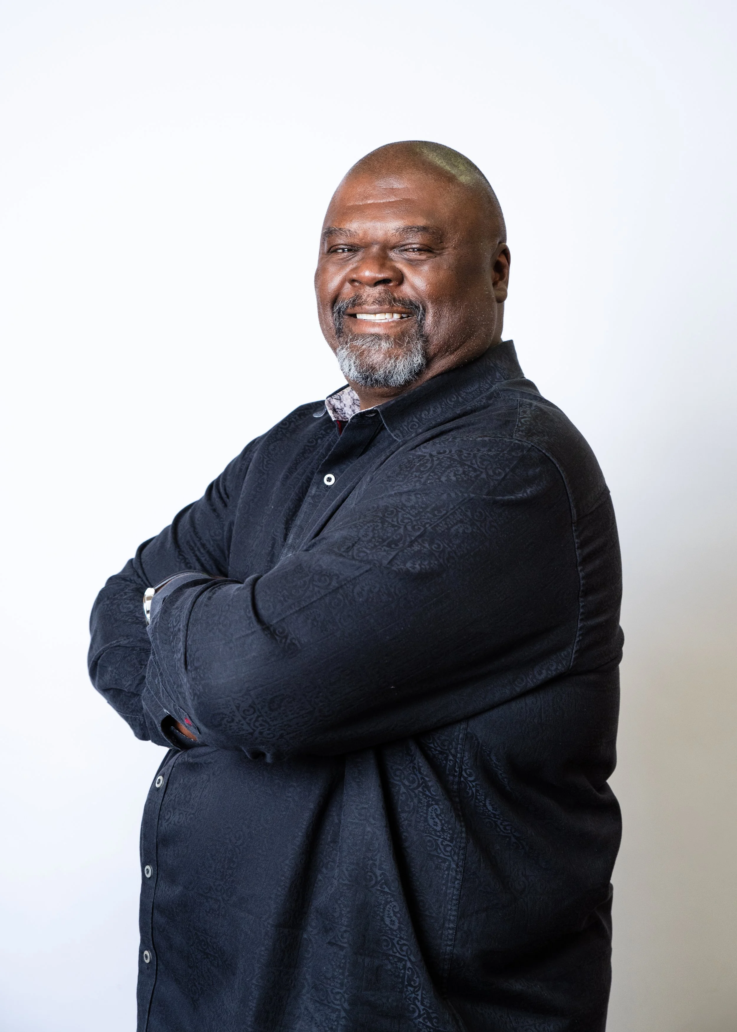A smiling Black man with a beard and short hair, wearing a black cardigan over a black shirt, standing against a peach-colored background.