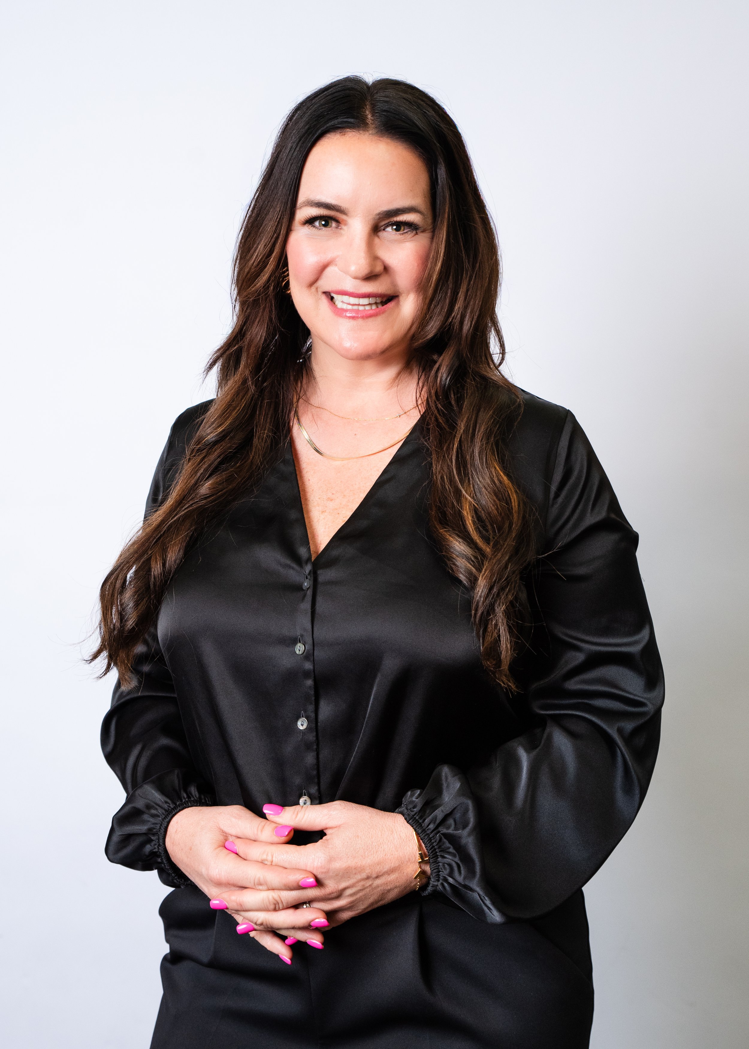 A woman with long brown hair, smiling confidently, wearing a black blazer over a black satin shirt, with blue jeans, standing against a warm gradient background.