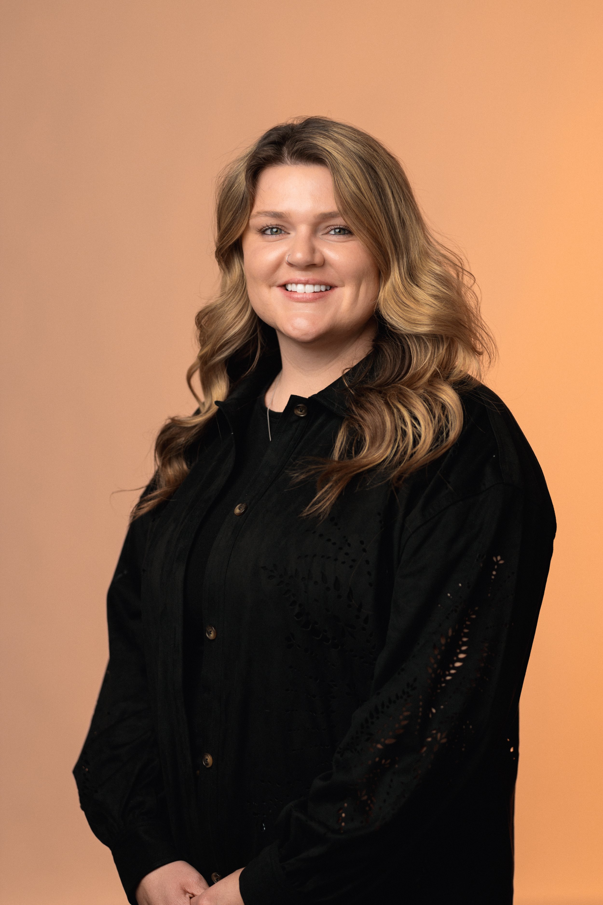 A young woman with wavy, light brown hair smiling, wearing a black shirt, against a gradient orange background.