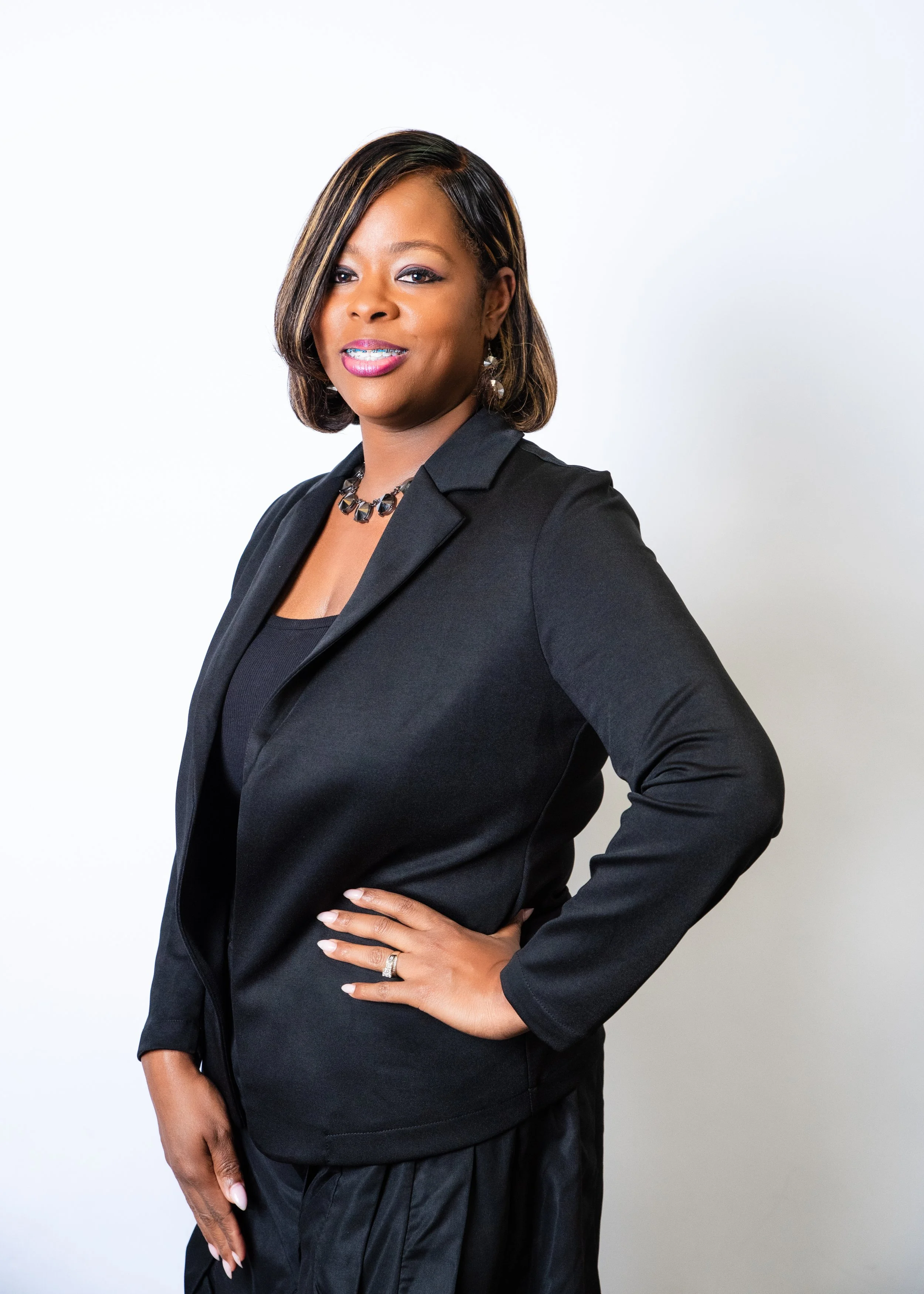 A woman with medium-dark skin and shoulder-length black hair, wearing a black blazer, a black and silver necklace, and earrings, standing against a beige background.