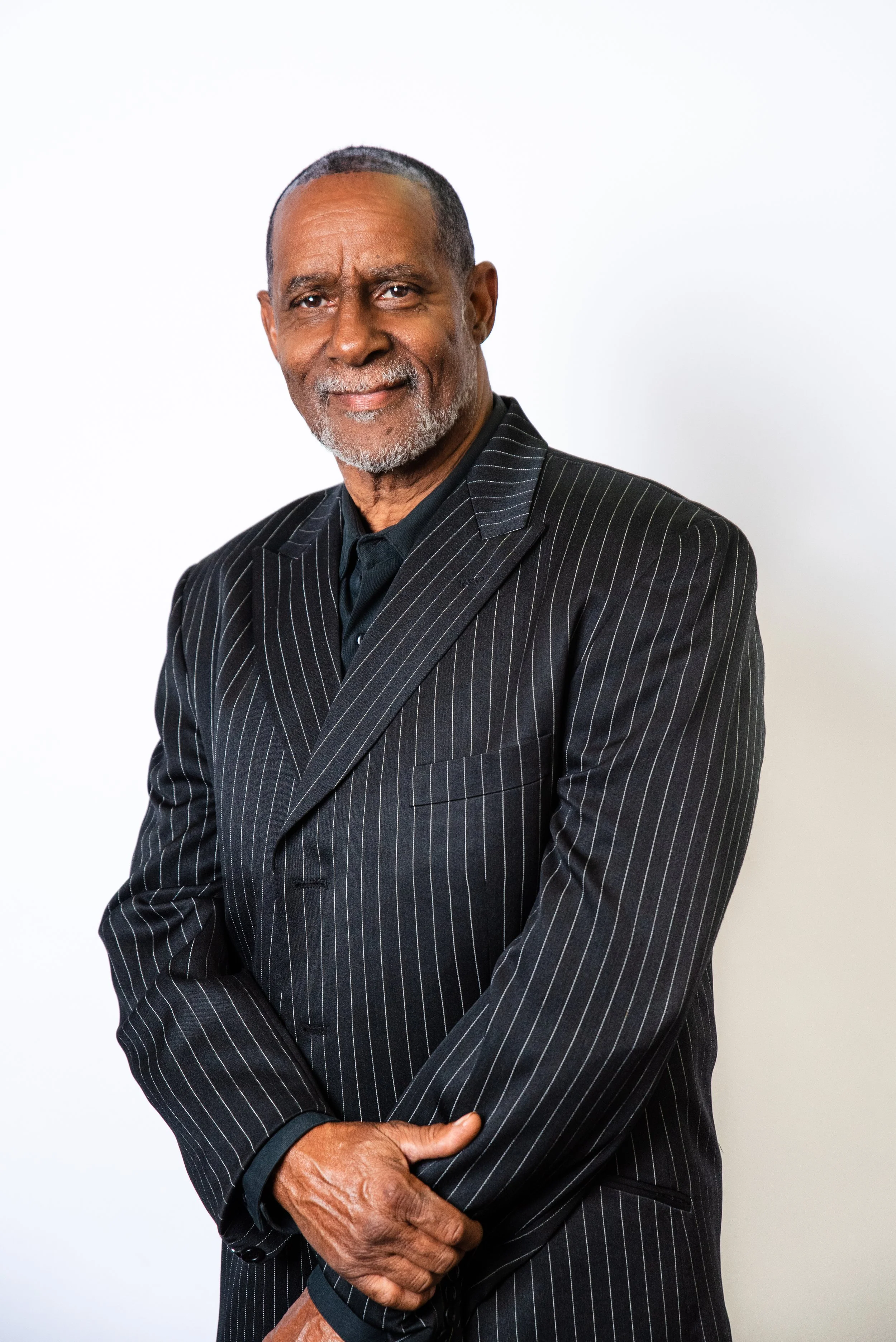 An older man with a gray beard and mustache, wearing a black suit and shirt, smiling against a peach-colored background.