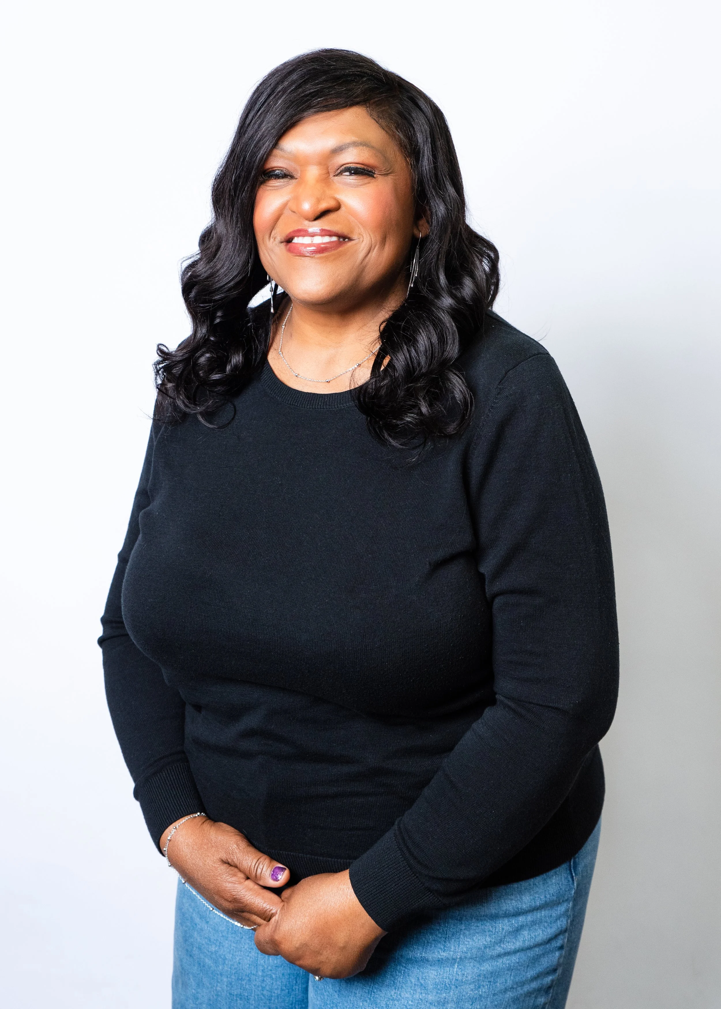 A smiling African-American woman in a black sweater and gold necklace standing against a peach background.