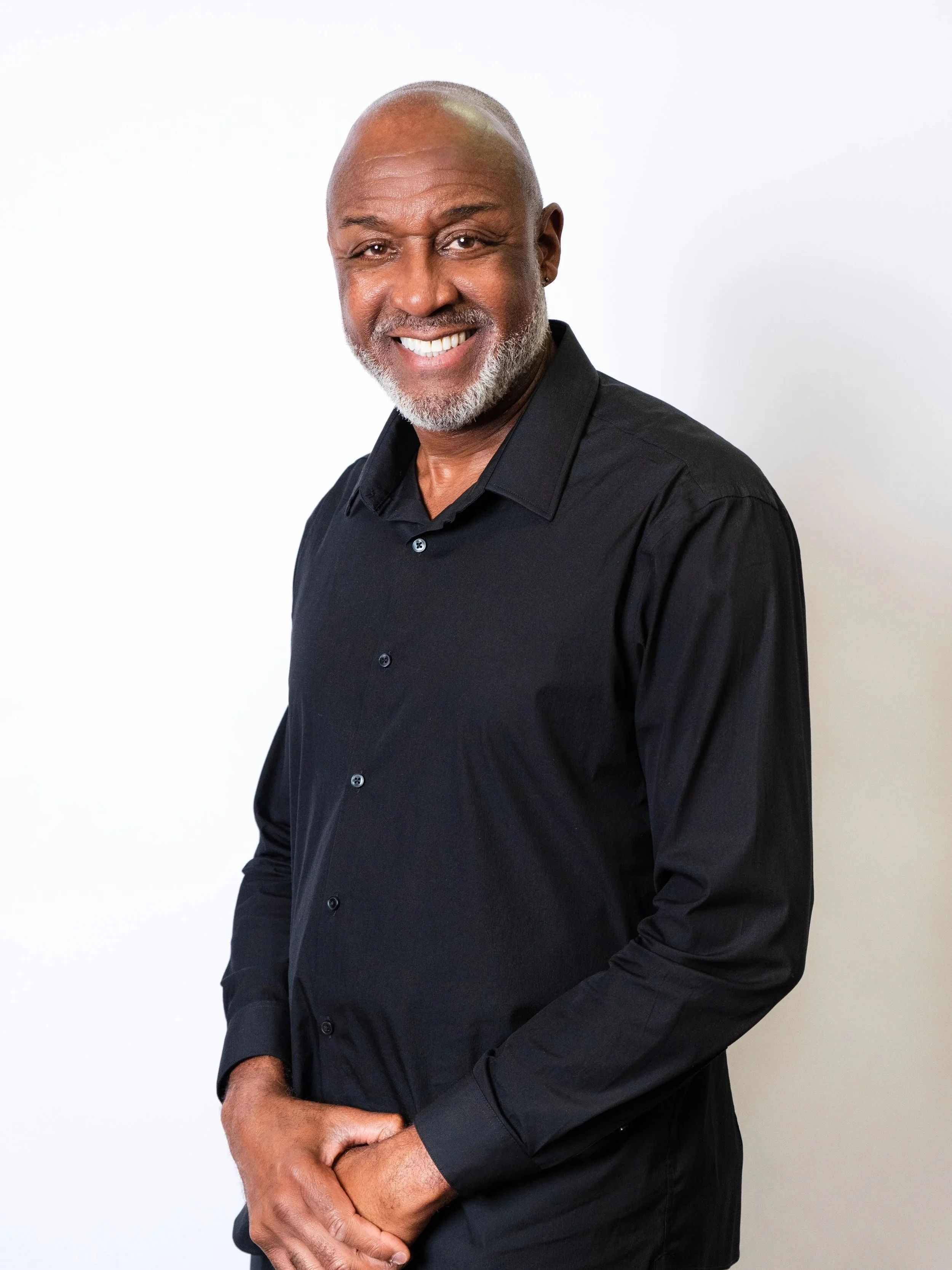 A smiling middle-aged man with a shaved head wearing a black button-up shirt, standing against a plain peach-colored background.