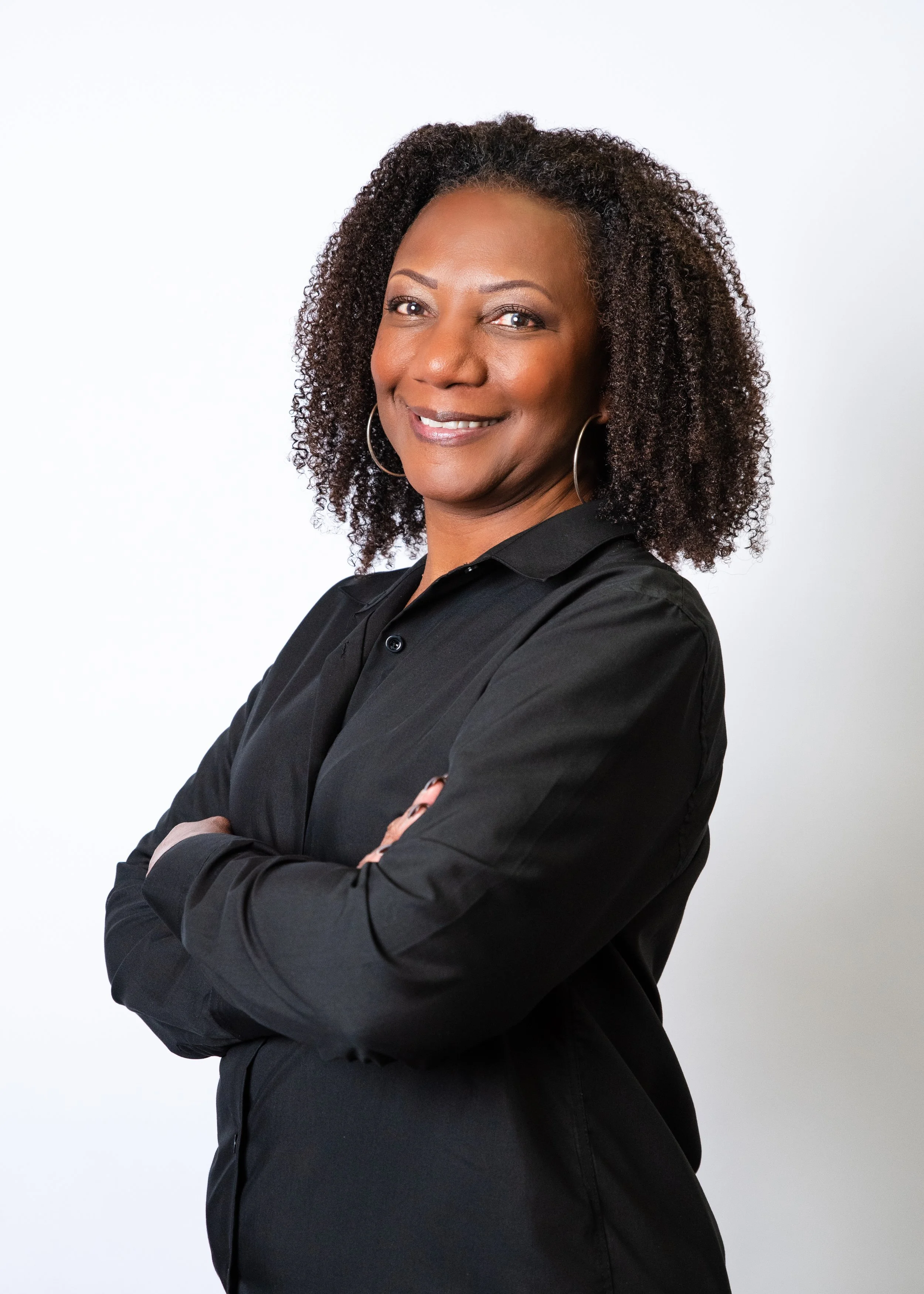 A woman with curly hair, wearing a black button-up shirt, smiling, with hoop earrings, against a plain, gradient background.