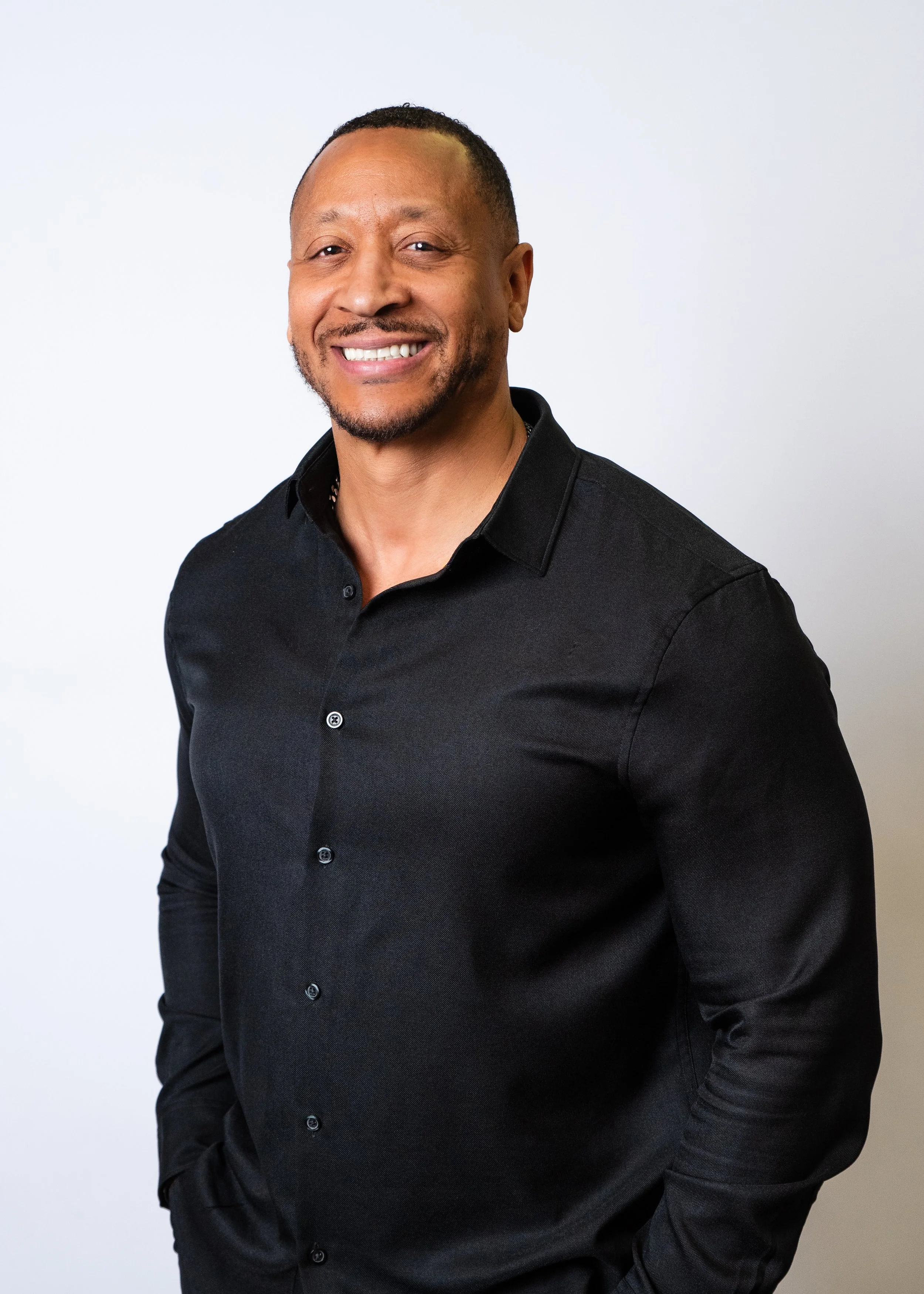 A smiling man in a black dress shirt standing against a plain, gradient orange background.