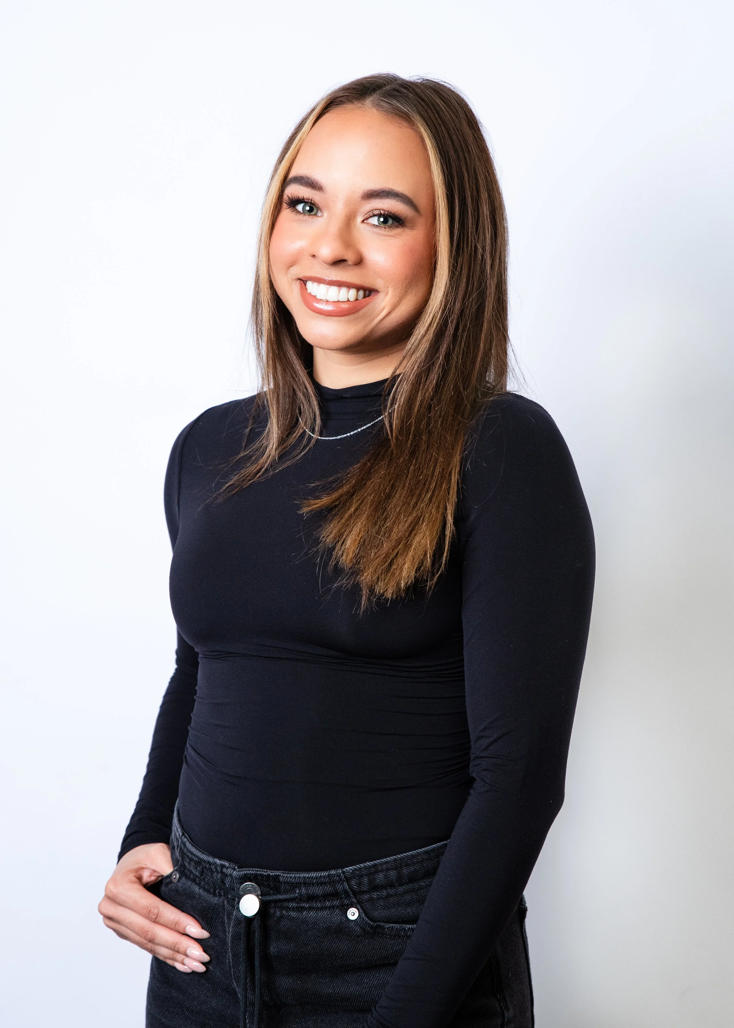A young woman with long, wavy dark hair and a bright smile, wearing a black top with a black jacket and jewelry, standing against a warm, gradient orange background.