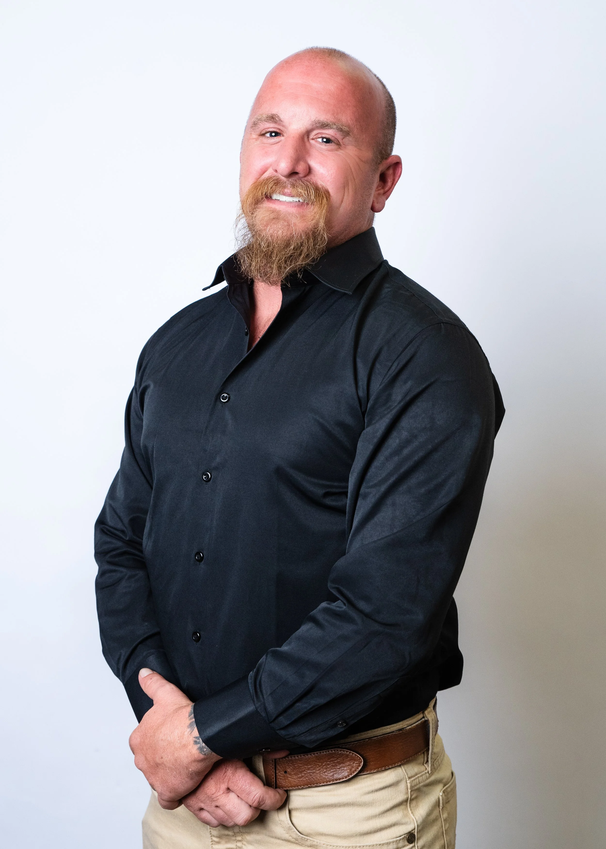 A man with a beard and bald head, wearing a black button-up shirt, standing against a gradient background with warm tones.