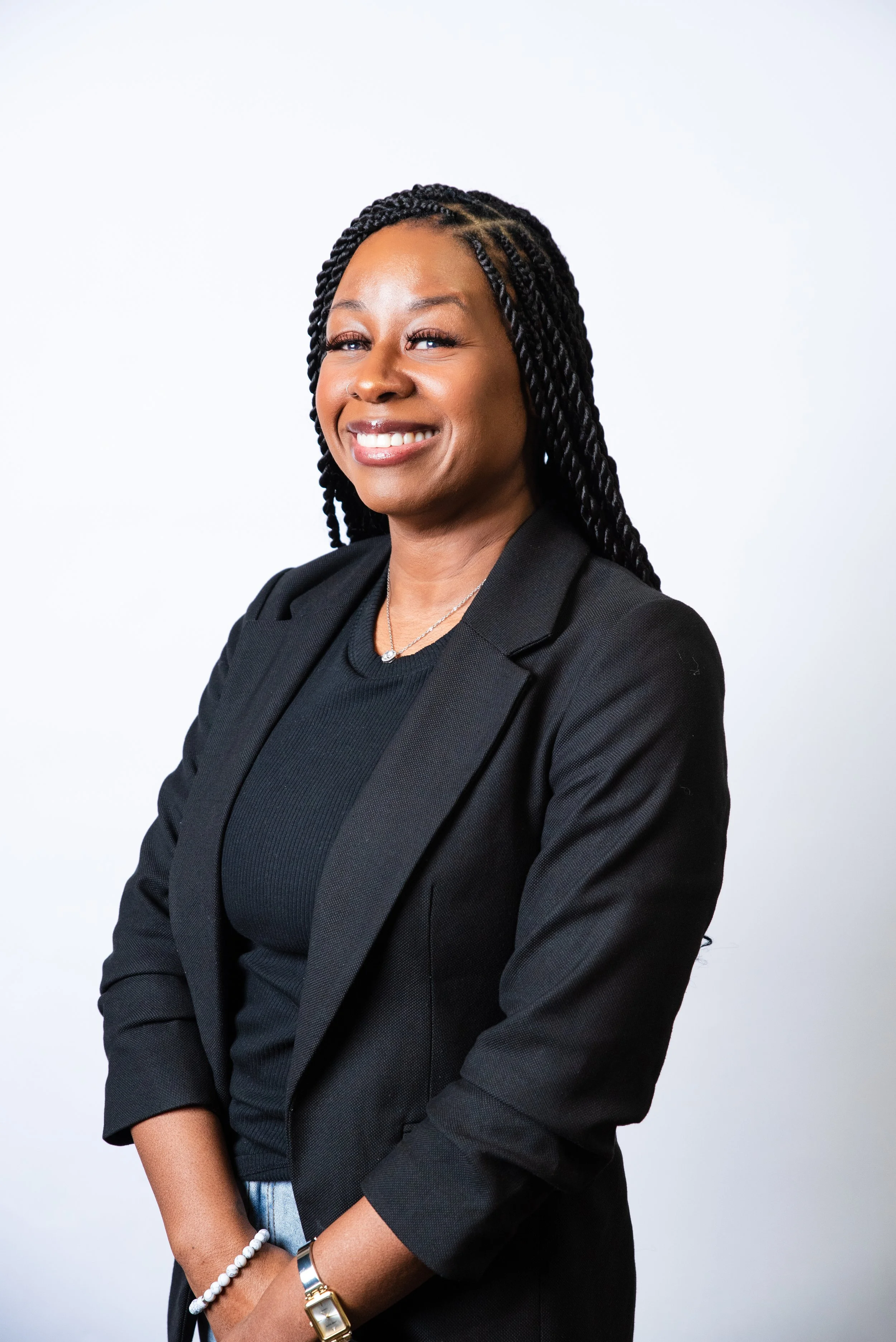 A woman with glasses and braided hair smiling, wearing a black ribbed long sleeve shirt, gold jewelry, and a watch, against a gradient peach background.