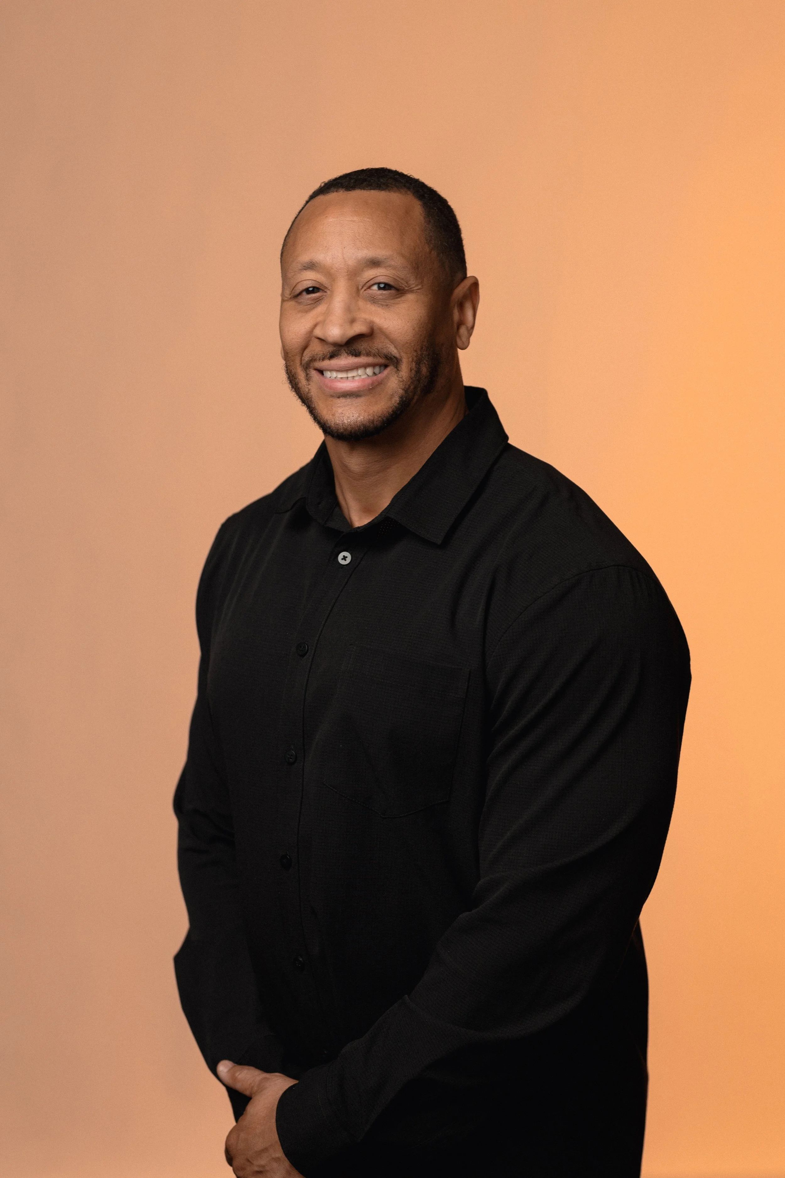 A smiling man in a black dress shirt standing against a plain, gradient orange background.