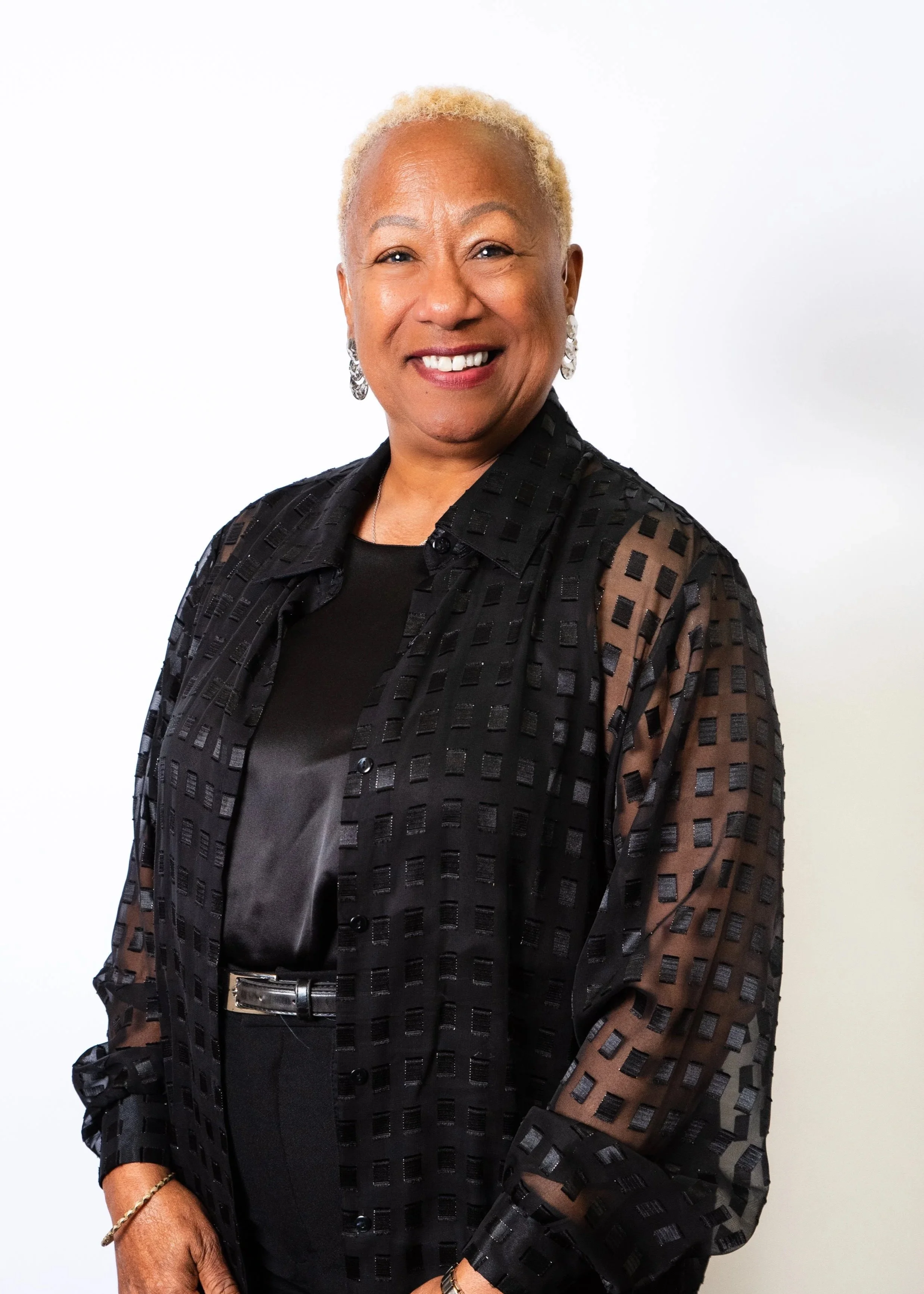 A portrait of an elderly woman with short, curly, reddish hair, wearing a black blazer and a matching top, with gold earrings, a necklace, and red lipstick, standing against a plain, peach-colored background, smiling.
