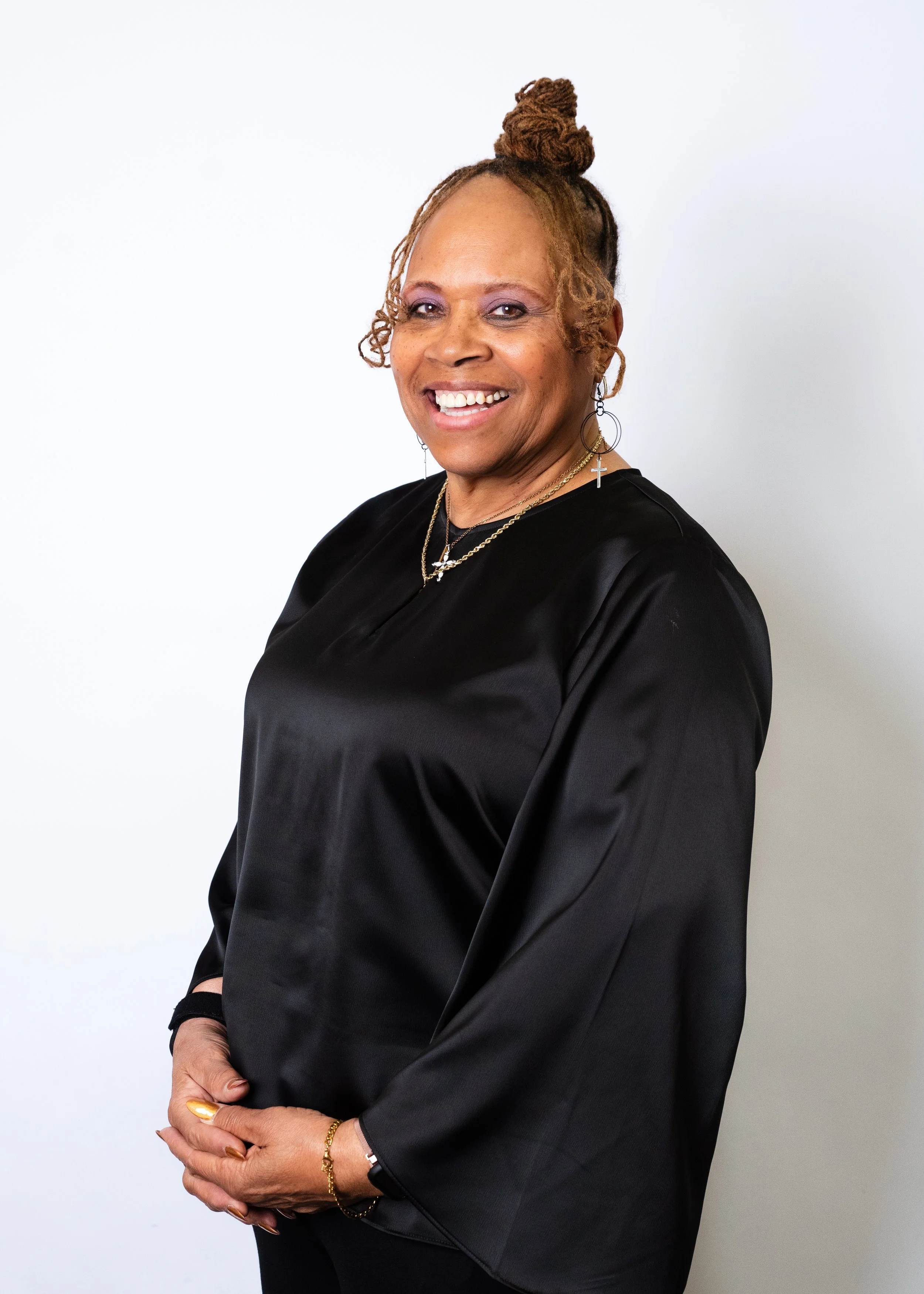 A woman with long red dreadlocks wearing a black long-sleeve top with ruffle details, standing against a warm beige background.