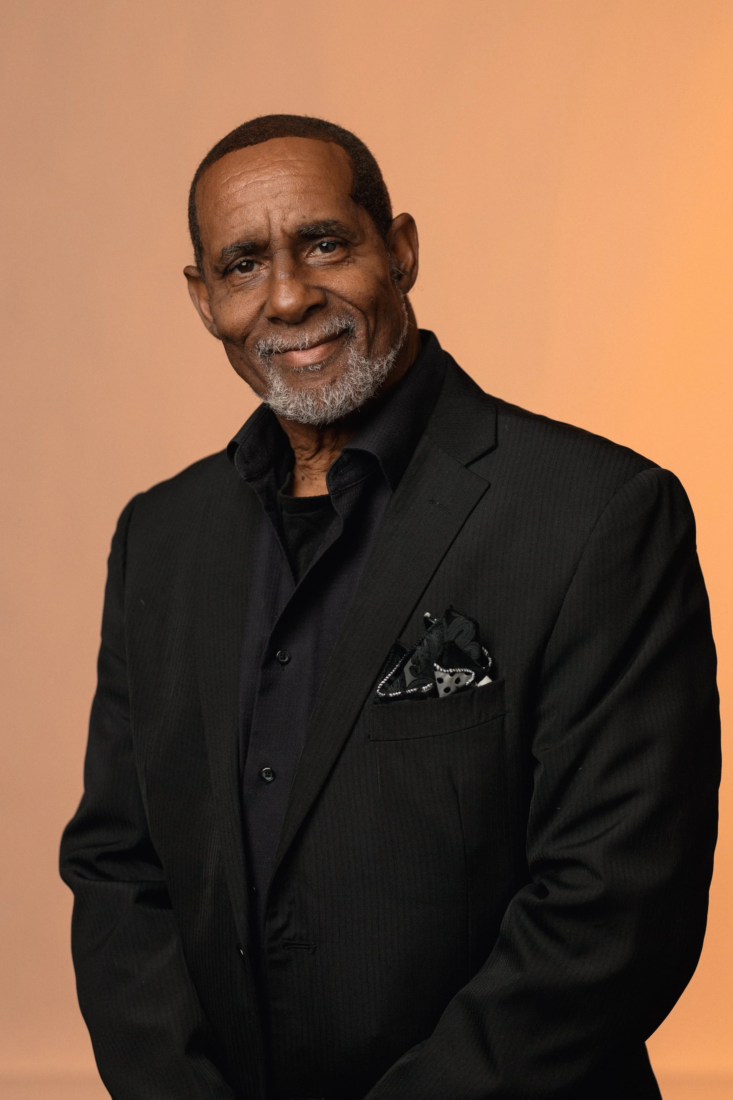 An older man with a gray beard and mustache, wearing a black suit and shirt, smiling against a peach-colored background.